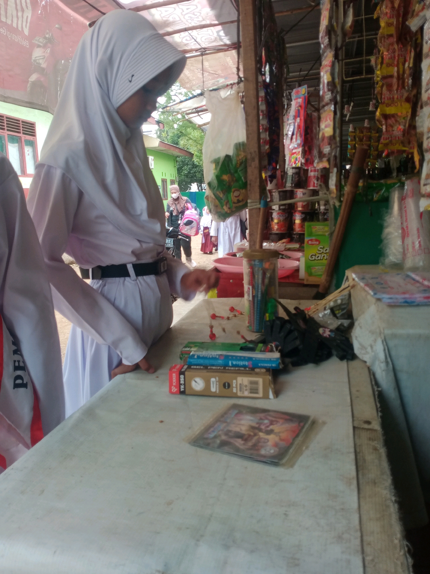 The image depicts an outdoor scene at what appears to be a small market stall in Bandar Lampung, Indonesia. A young person wearing a white hijab and school uniform stands to the left, leaning over a table. The uniform consists of a white long-sleeved shirt and a white pleated skirt, cinched with a black belt. Another person in a similar uniform is partially visible to the far left. The table in the foreground is covered with a light-colored, textured cloth and displays several items including stacks of boxes containing what appear to be stationery or small packaged goods, and a CD or DVD case.
Behind the table, the market stall is visible with various goods hanging and displayed. These include rows of colorful packaged snacks and other products. A wooden pole supports the stall's canopy. In the background, more people are seen, including a woman in a headscarf wearing a mask and carrying a pink bag, and other individuals in what seems to be a school environment, suggesting the market is located near a school. The general environment appears to be a typical roadside stall in a tropical climate. The lighting suggests it is daytime.