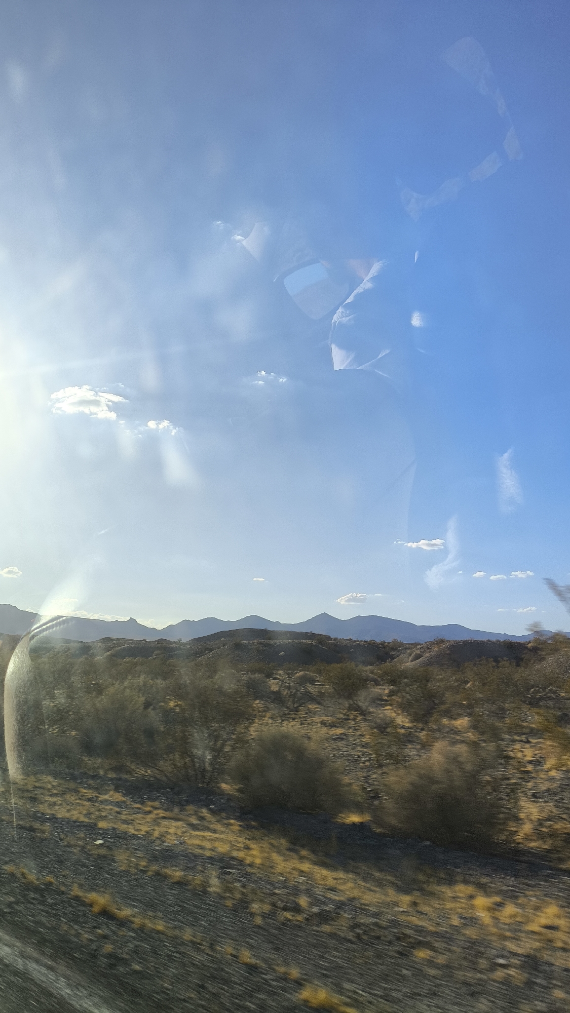 The image captures a desert landscape under a bright, clear blue sky with scattered wispy clouds. The sun is visible to the left, casting strong rays across the scene. In the foreground, a textured, rocky ground with sparse, dry vegetation is depicted, suggesting a desert environment. Low, rounded hills form the middle ground, sparsely covered with shrubs and trees. In the distance, a range of larger mountains stretches across the horizon.
Reflections are present on what appears to be a window, showing a person's head and shoulders, likely the photographer, and a portion of what could be a vehicle's interior, such as a side mirror. The overall impression is of a scenic drive through a dry, arid region. The location is identified as Shoshone, United States, which is consistent with a desert landscape.