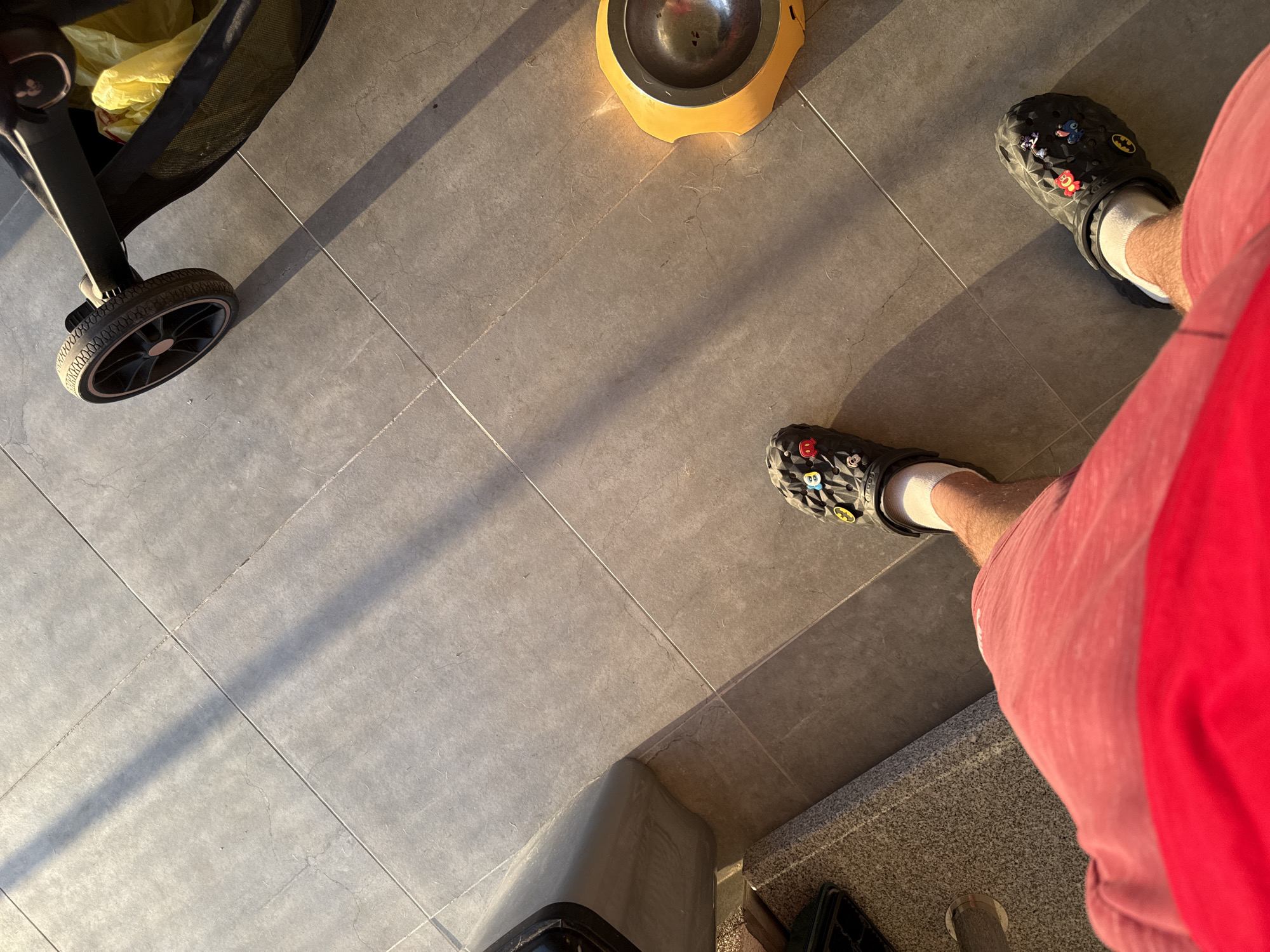 The image captures a ground-level perspective, likely taken indoors on a tiled floor. A stroller with a black frame and filled with a yellow plastic bag is located on the upper left. A yellow dog or cat bowl is visible near the upper center of the image. To the right, a person is seen wearing pink shorts and black Crocs with assorted charms, accompanied by white socks. The individual's legs are captured from the knees down. Based on the information provided, the location is Sofia, Bulgaria.