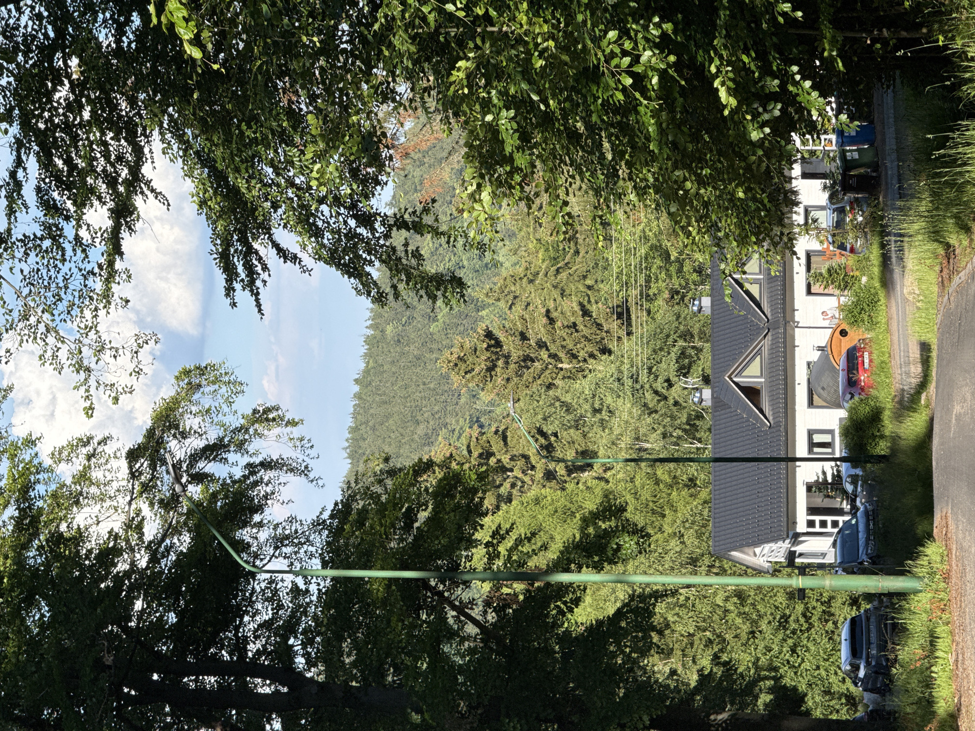The image depicts a residential building nestled amongst lush green trees with a backdrop of a forested mountain range. A paved driveway leads to the house, where several cars are parked. The house itself is a white structure with a dark tiled roof and several dormer windows. Two green lampposts are visible on the property, one on the left and another further down the driveway. The sky is a clear blue with scattered white clouds. The overall scene suggests a peaceful, secluded location, likely in a rural or semi-rural area. The image appears to be taken on a sunny day, with ample natural light illuminating the scene. Based on visual cues, this could be in or around Szklarska Poręba, Poland, known for its natural beauty and mountainous terrain.