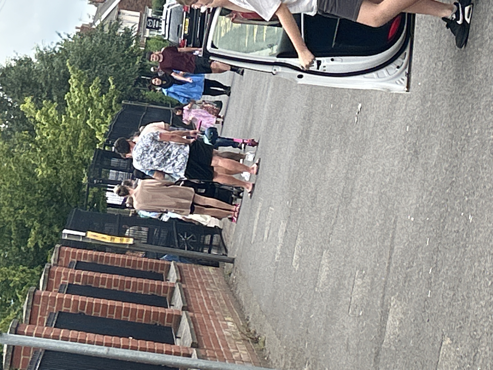 An outdoor scene depicts a group of people walking along a paved walkway bordered by a brick wall and a metal fence. In the foreground, the open door of a white car is visible. Further down the walkway, a man in a patterned shirt and dark shorts is seen walking, followed by other individuals. Some people are carrying bags, and one person is pushing a stroller. A pink scooter is parked on the walkway. In the background, trees and houses are visible, suggesting an urban or suburban setting. The lighting indicates it is daytime. The location is identified as Boston, United Kingdom.