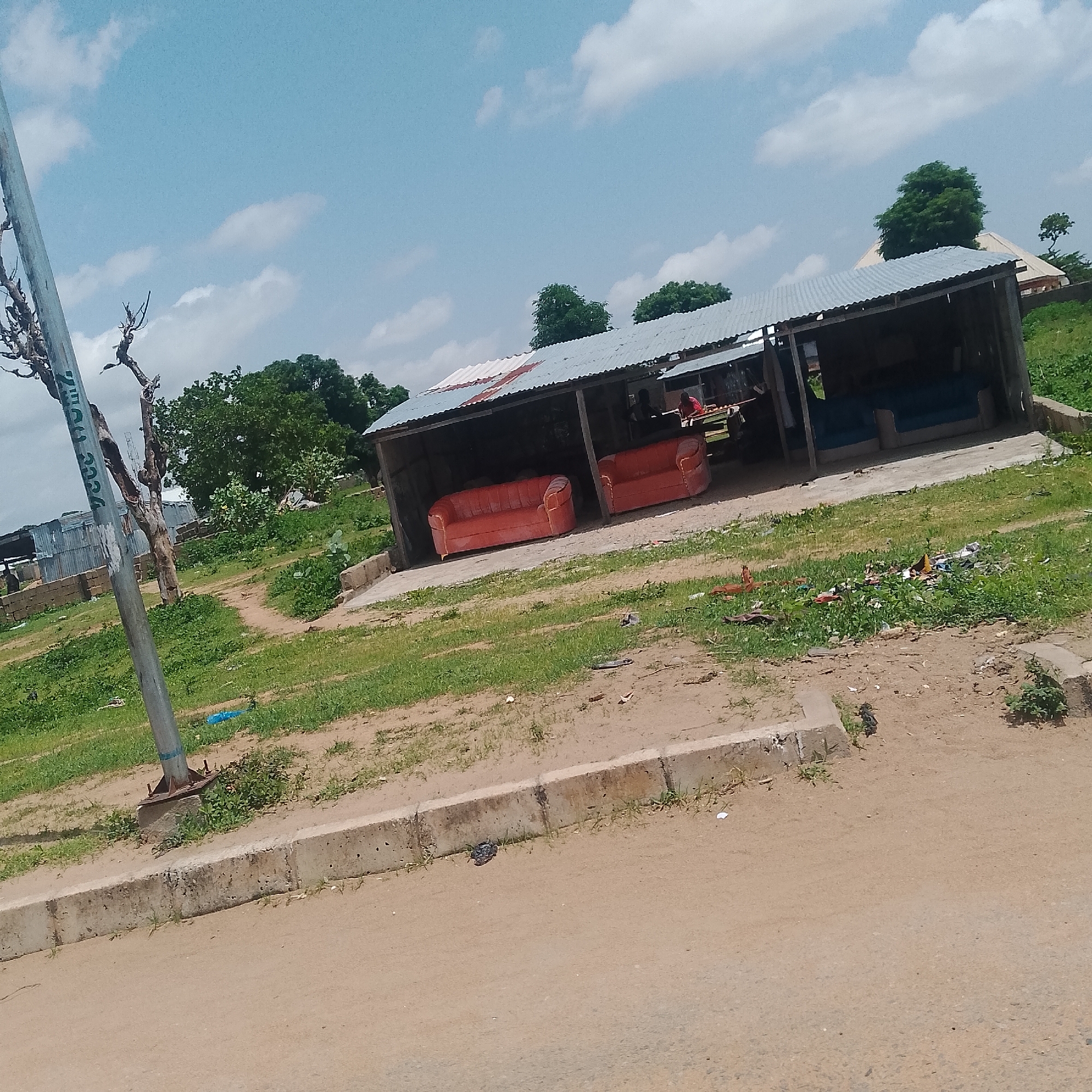 The image depicts an outdoor scene in Jalingo, Nigeria. In the foreground, a sandy dirt road borders a concrete curb. Beyond the curb, an area of dry, patchy grass is visible, with some areas of compacted dirt. Further back, under a corrugated metal roof supported by wooden posts, two bright orange couches are placed on a concrete slab. To the right of the couches, another section of the structure reveals what appears to be a blue couch or mattress.
To the left of the sheltered area, a tall, weathered utility pole stands, showing some graffiti or markings. Behind the pole, a dead, gnarled tree is present. Beyond the structure and the dead tree, a line of trees and foliage forms a backdrop against a light blue sky with scattered white clouds. In the distance, a few buildings with metallic roofs are visible, suggesting a semi-urban or fringe-of-city setting. Scattered on the ground in front of the sheltered area, and to the right of it, is a collection of debris, including plastic bags and other refuse.
The overall impression is that of a commercial or informal gathering space, perhaps a roadside shop or rest area, in a somewhat arid environment. The scene is illuminated by natural daylight, indicating it was taken during the day. There are no people prominently featured in the image, though silhouettes suggesting human presence can be inferred within the sheltered structure. The weather appears to be clear and partly cloudy.