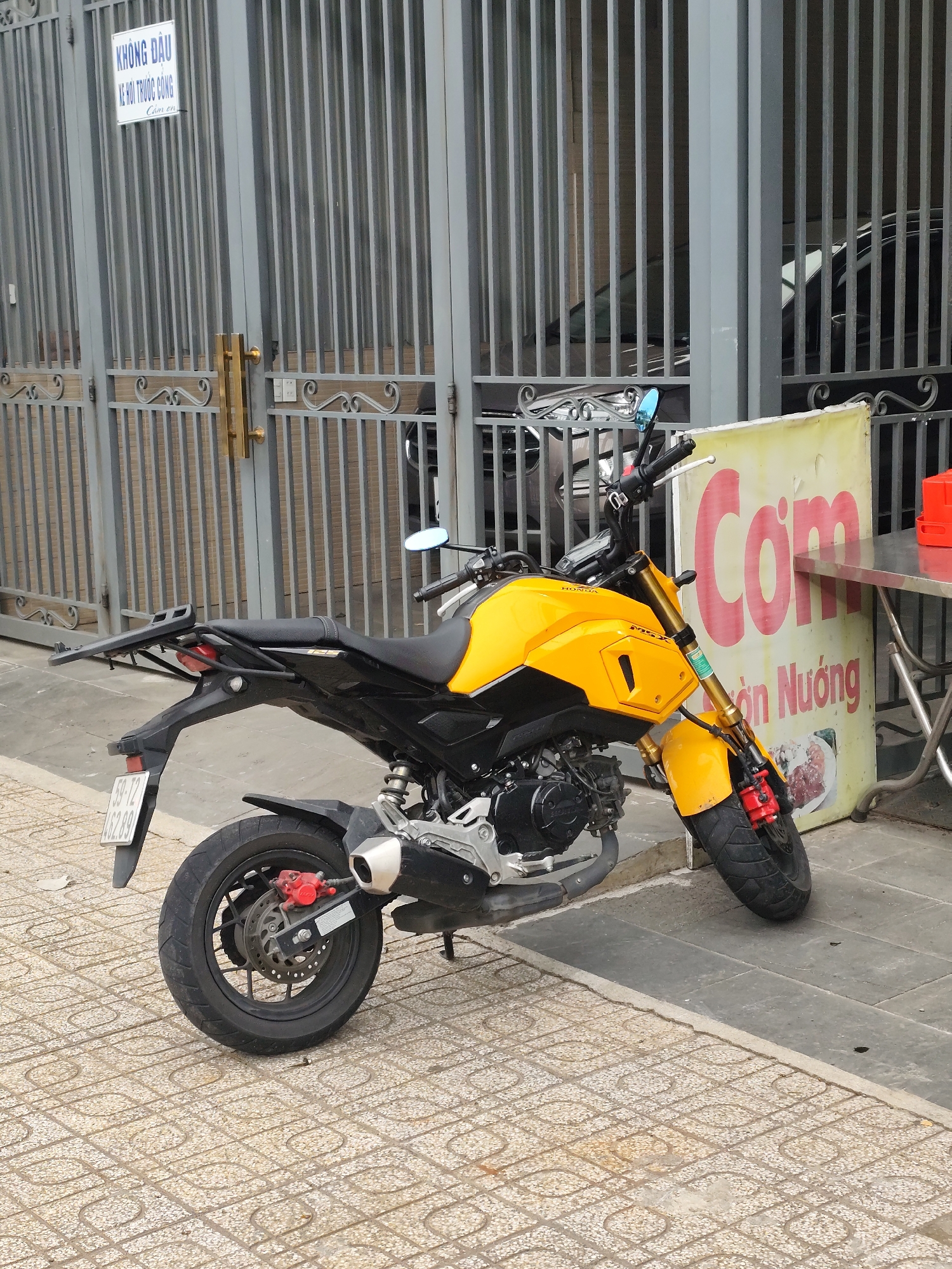 A yellow and black Honda Grom motorcycle is parked on a sidewalk in front of a grey metal gate. Behind the gate, the front end of a black car is visible. To the right of the motorcycle is a sign with Vietnamese text and a small metal table. The motorcycle has a license plate with the number "59-T2 462.89". The gate has a sign that reads "KHÔNG ĐẬU XE KHỎI THUỐC CỘNG" in Vietnamese. The sidewalk is paved with patterned tiles. Lâm Viên - Đà Lạt, Vietnam is implied from the Vietnamese text on the signs.