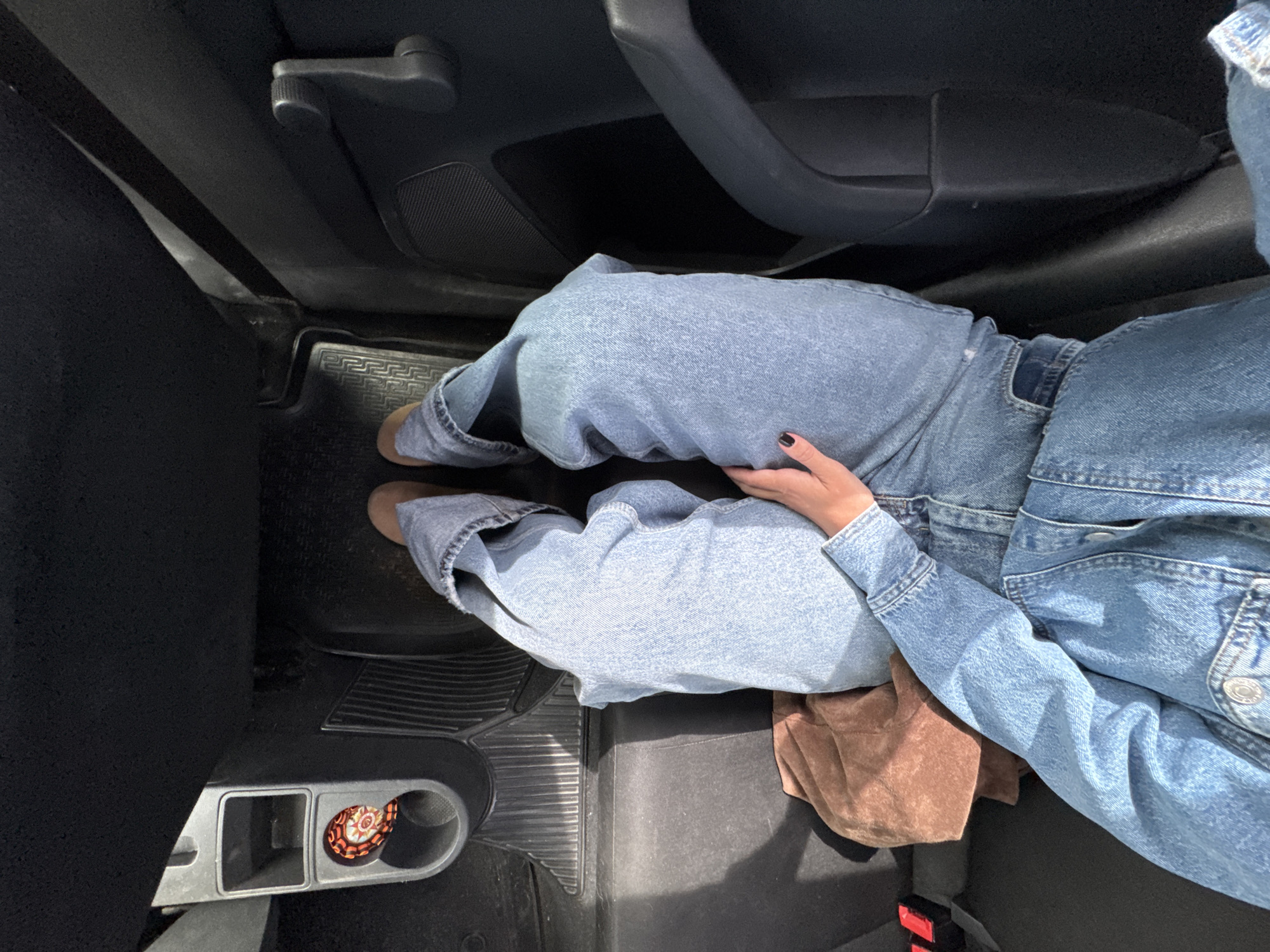 The image shows a person's legs in light blue denim jeans and a denim jacket, sitting in the back seat of a car. A brown fabric item, possibly a scarf or bag, is to the person's left. The car's interior is dark gray and black. The view is from above, looking down at the person's legs and lap. This image was taken on August 4, 2023, at 12:16 PM, in Chaikovsky, Russia.