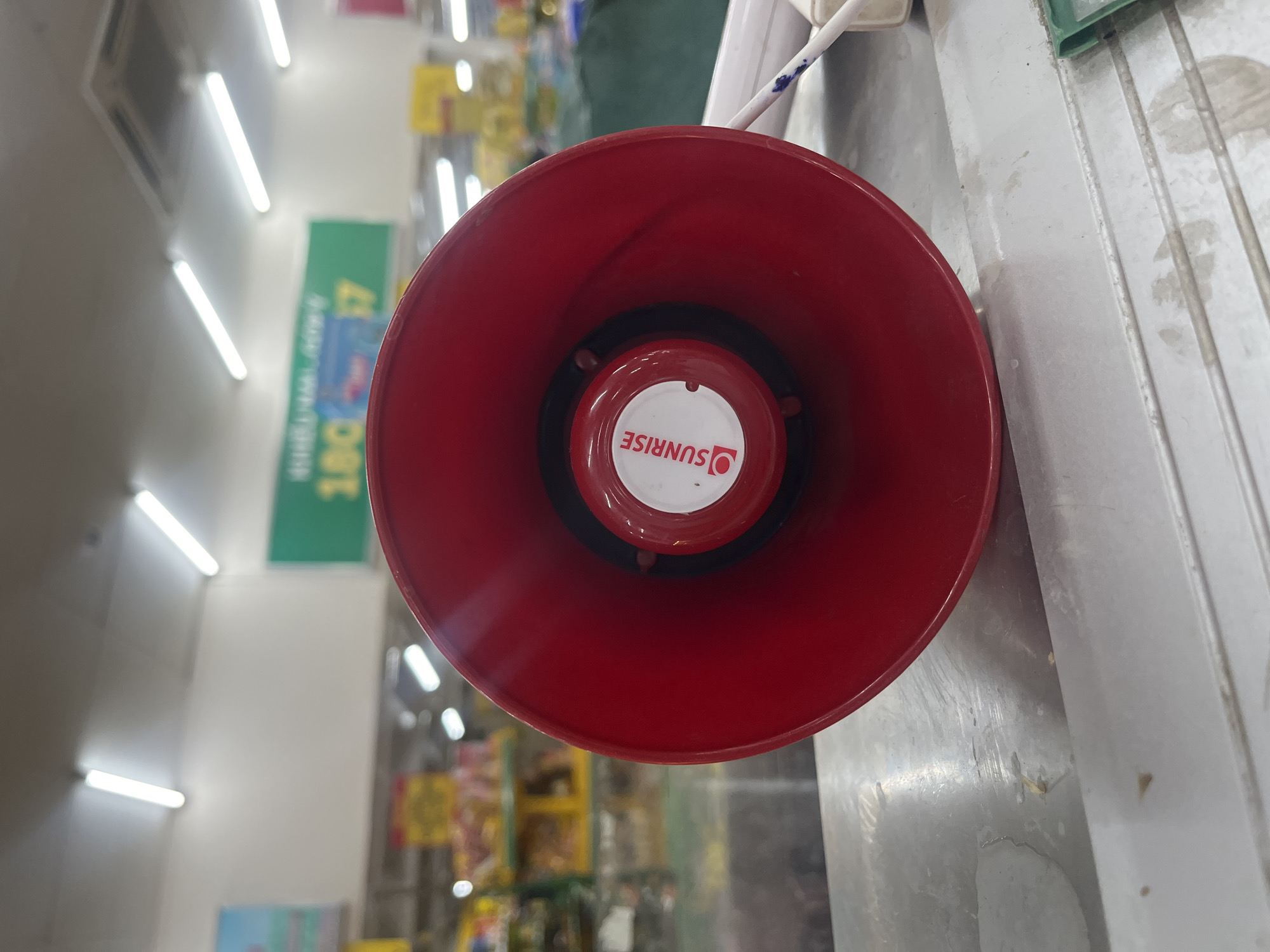 The image shows a close-up of a red horn speaker with a white circular sticker that has the logo "SUNRISE" printed on it. The speaker is positioned on a metal surface. In the background, a blurred retail environment is visible, with fluorescent lighting on the ceiling and shelves stocked with goods. A green sign with white text is also visible in the background. The overall impression is that this image was taken in a store or commercial establishment in Bac Nha Trang Ward, Vietnam.