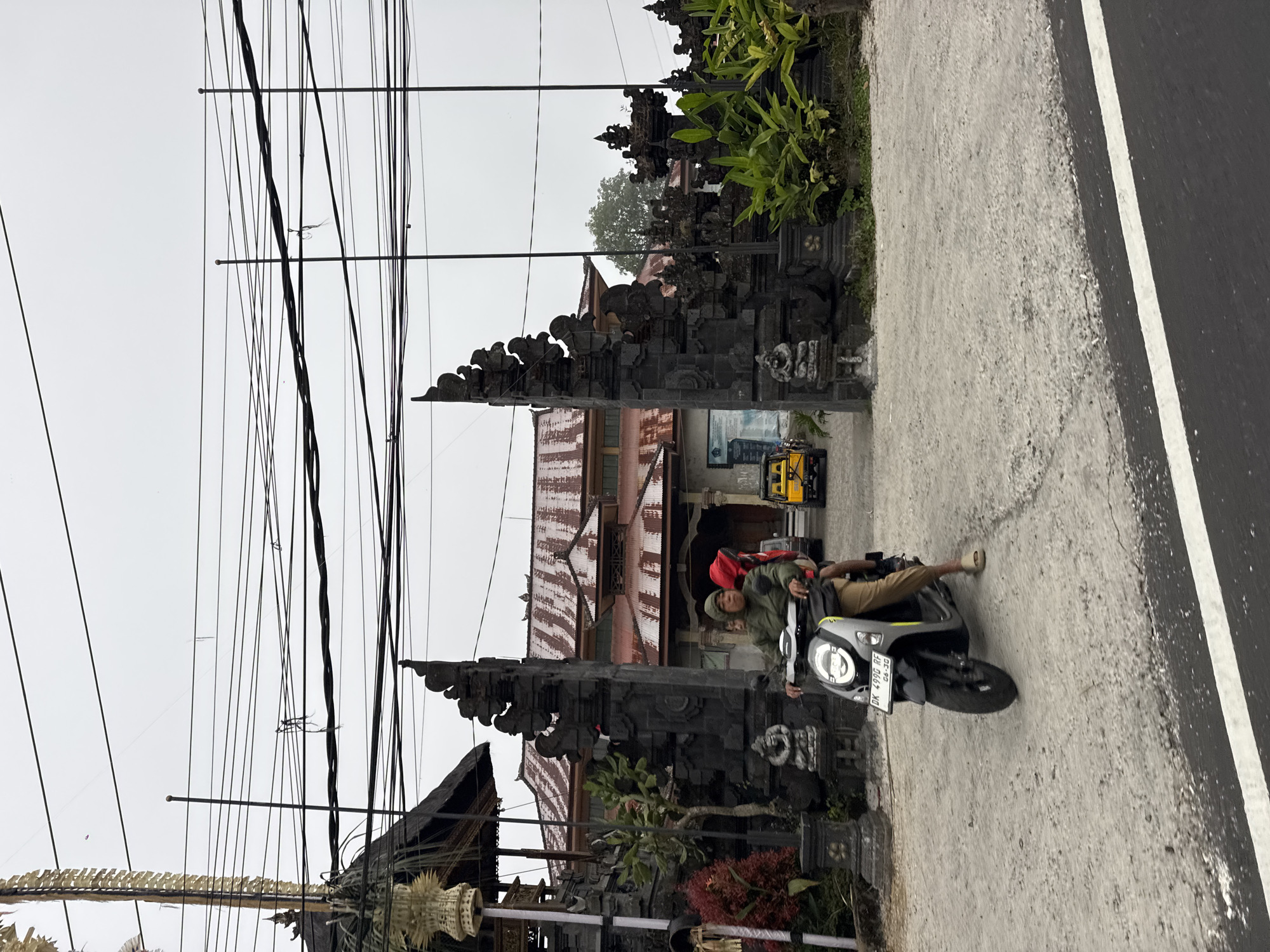 A person rides a scooter past a stone gate and traditional Balinese buildings. The scooter, a silver Honda, has a license plate that reads "DK 499G RF 06-20". The rider wears a green jacket and a cap, with a red backpack visible. They are riding on a paved road with a white line marking. Behind the gate, which is intricately carved from dark stone, are buildings with corrugated metal roofs, some appearing aged. Lush green plants grow around the gate and near the buildings. The sky is overcast, suggesting a cloudy or possibly rainy day. The scene appears to be a street in Kintamani, Indonesia, characterized by the distinct architecture and vegetation. The overall atmosphere is that of a typical day in a rural or semi-urban Indonesian setting.