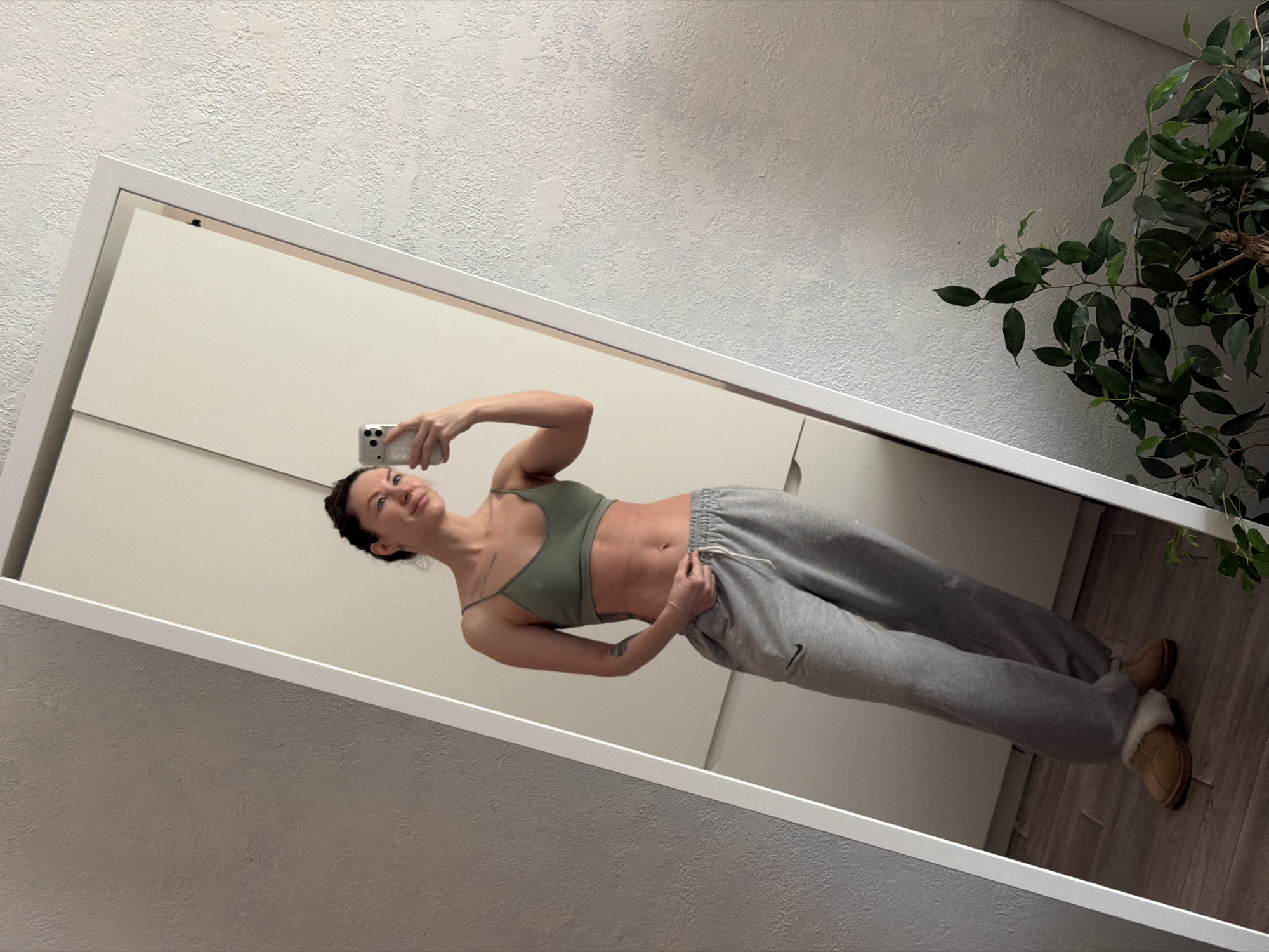 A woman stands in front of a large mirror, holding a smartphone in her right hand as if taking a selfie. The reflection in the mirror reveals her wearing a green sports bra and gray sweatpants with a Nike logo on the left leg. She's wearing tan-colored slippers with fur lining. Her hair is pulled back into a bun, and she is looking at the phone. The environment consists of a white wall with textured finish and a plant with green leaves visible on the right edge of the mirror. Based on available information, no details indicating the location to be in Perm, Russia, are present.