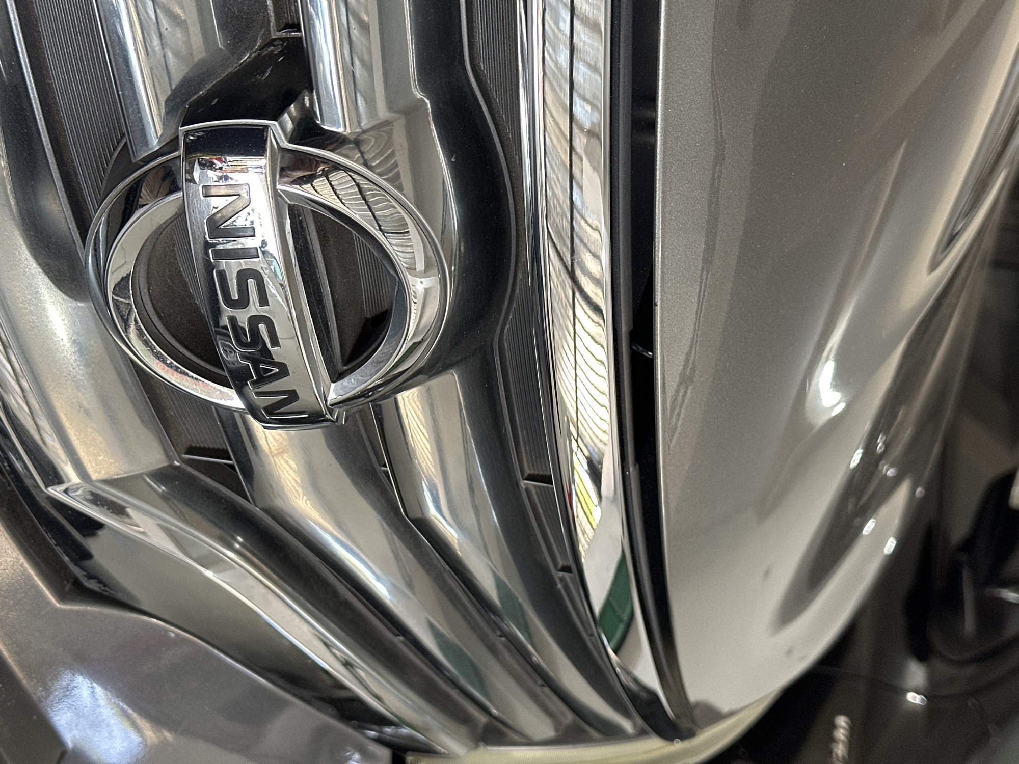The image is a close-up, eye-level, medium shot of the front grille of a gray Nissan vehicle. The Nissan logo is prominently displayed in the center, appearing as a chrome emblem within a circular frame. The grille itself is made of chrome and features horizontal bars. The background is blurred, showing the car's hood and a hint of another dark vehicle behind it, possibly in a parking lot or garage setting. The lighting suggests an indoor or shaded environment. The location, Bulakrejo, Indonesia, is noted but not visually evident within the frame.