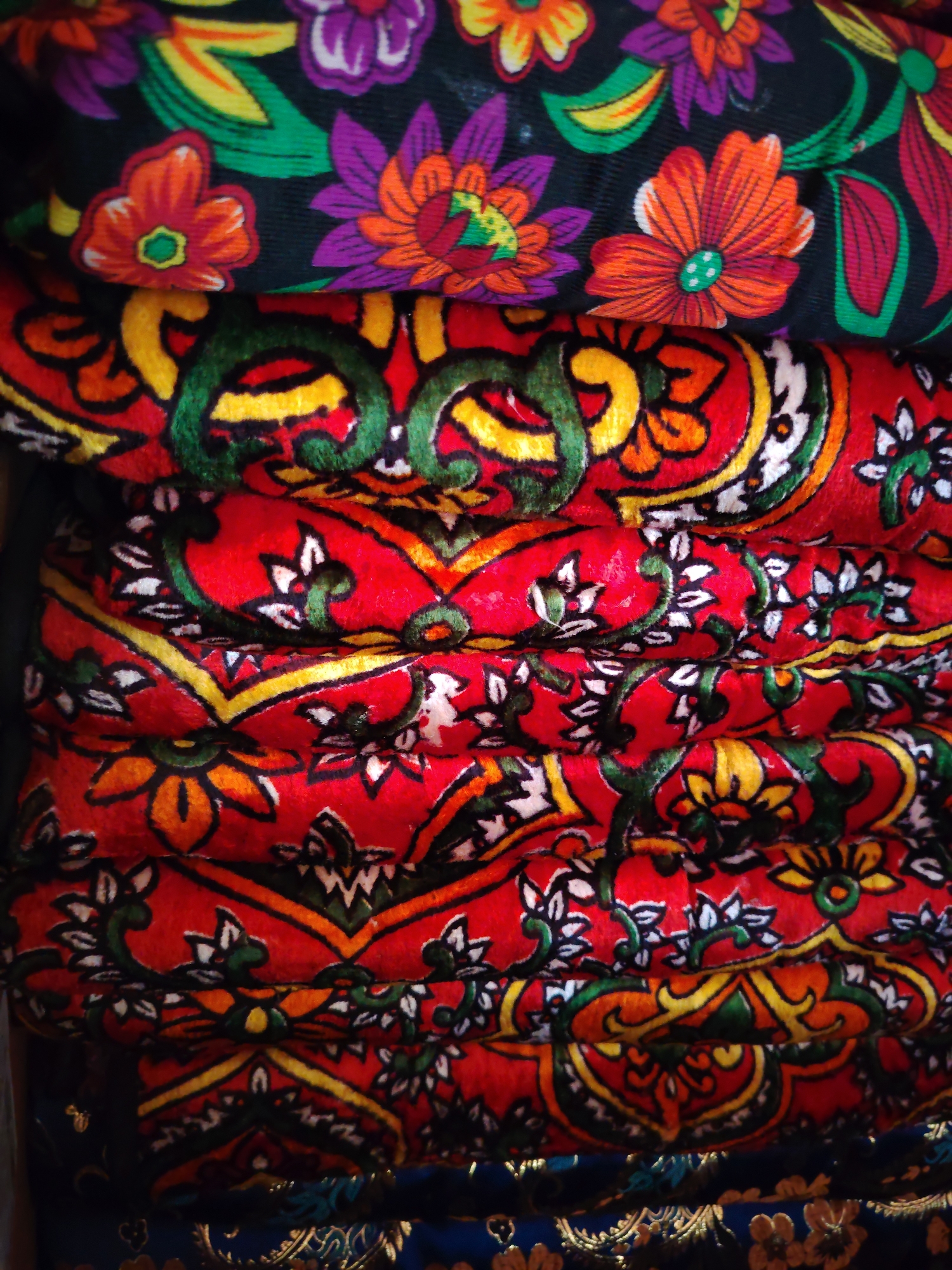 A stack of vibrantly colored, patterned textiles is displayed outdoors, possibly in a marketplace or a shop in Qayqi, Uzbekistan. The textiles are folded on top of each other, creating a layered effect. The topmost fabric features a dark background with a floral pattern of orange, purple, and green flowers, along with green leaves. Beneath this, several layers of red fabric are visible, each adorned with intricate paisley-like designs in yellow, green, and white. The lower portion of the stack shows a dark blue fabric with a metallic gold pattern, suggesting a different type of textile or an item beneath the red ones. The lighting suggests it is daytime, and the environment appears to be a setting where such textiles would be sold or displayed. There are no people visible in the image.
