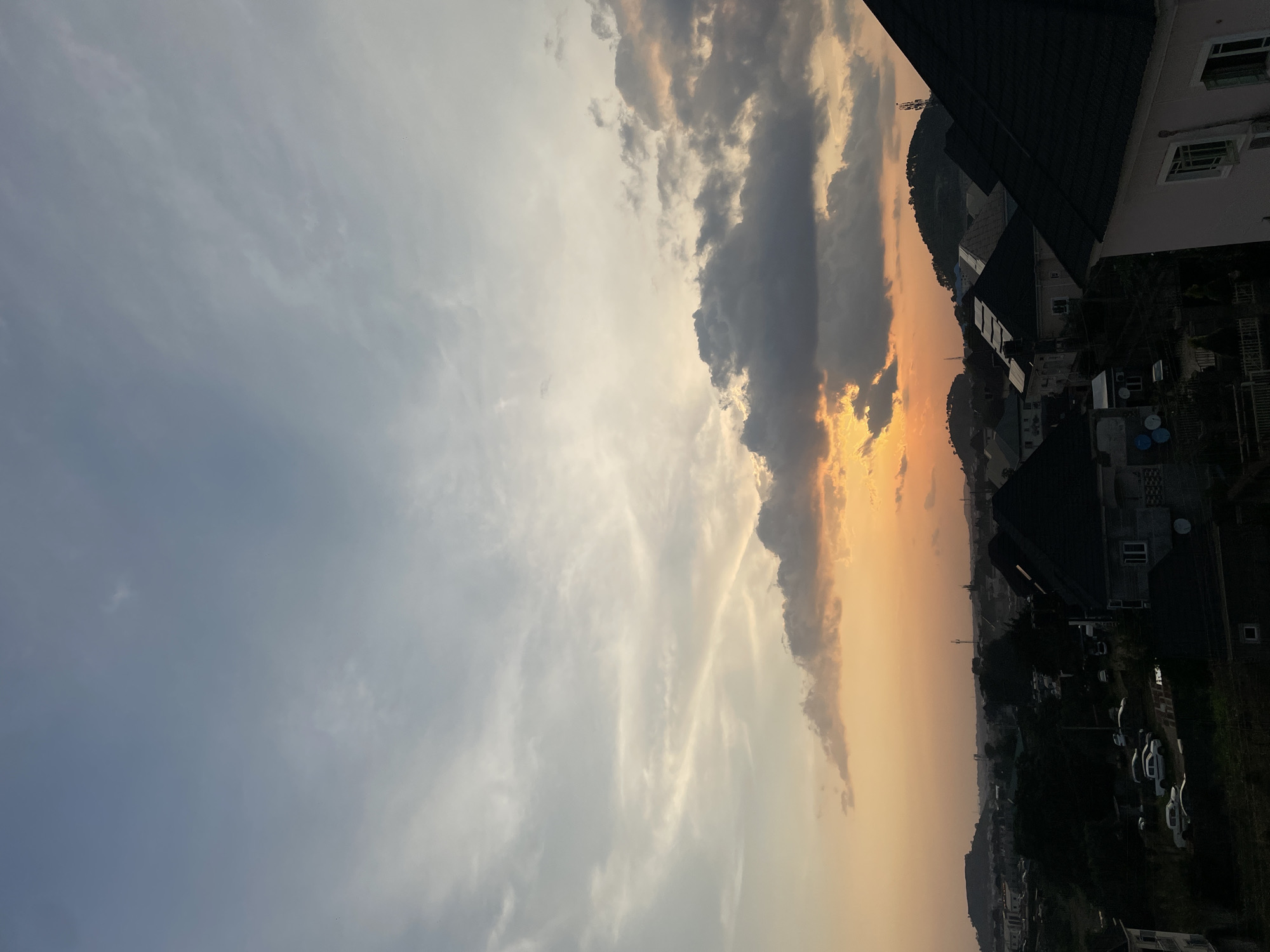 The image depicts a sunset scene over a residential area in Abuja, Nigeria. The sky is a blend of soft orange and gray hues, with scattered clouds. In the foreground, several houses with dark pitched roofs are visible, some with lights on. A street with parked cars is seen in the middle ground, bordered by trees. Distant hills and buildings form the background, with a tower visible on one of the hills. The overall impression is of a quiet evening in a suburban part of the city.
