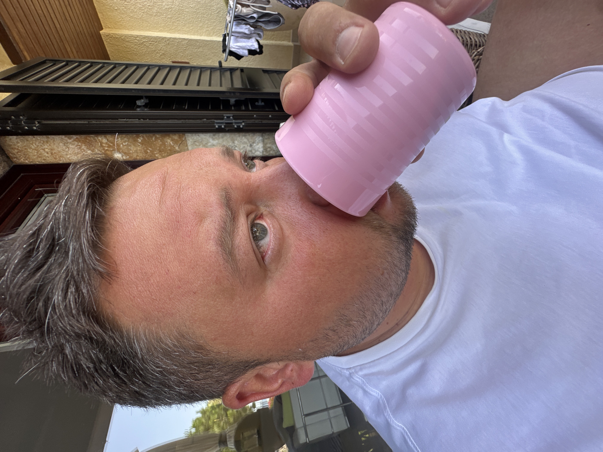 A close-up, eye-level, outdoor, daytime shot shows a man with short, dark hair with gray streaks, and stubble, drinking from a ribbed, pink cup. He is wearing a white t-shirt. The man's right eye is visible and appears to be blue-green. The background is out of focus but suggests a residential or resort setting with textured walls, dark-colored shutters, and a clothesline with laundry. The lighting indicates it is a sunny day. The man is holding the cup with his right hand. The location is identified as Sant Feliu de Guíxols, Spain.