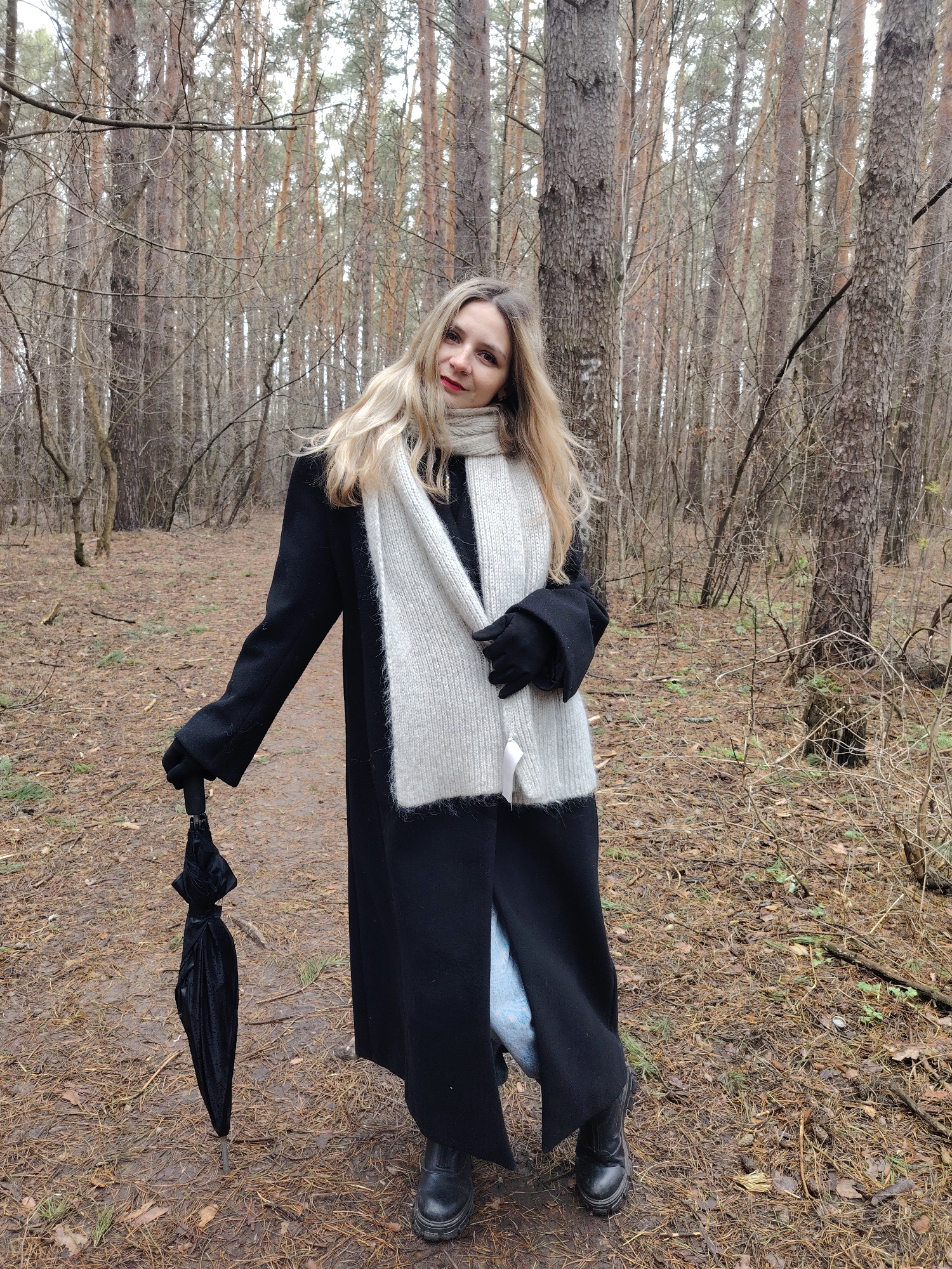 A woman is standing in a forest. She is wearing a long black coat, a light-colored scarf, black gloves, and black boots. She is holding a closed black umbrella in her left hand. The forest has tall, bare trees and a ground covered in pine needles and twigs. The woman has long blonde hair and red lipstick. The location is Chaikovsky, Russia.
