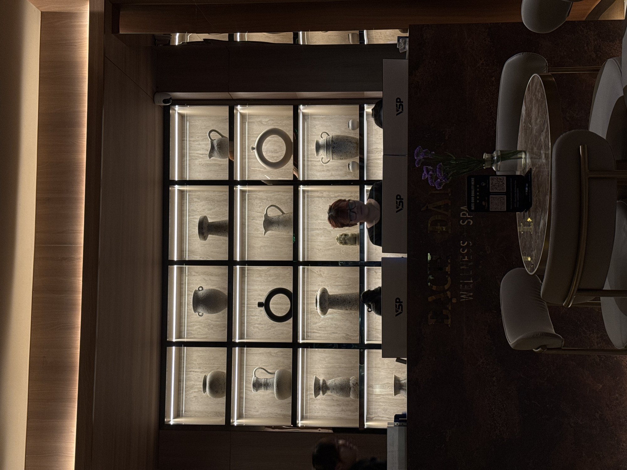 The image is of a spa or wellness center. In the background is a wall of square compartments, each containing a decorative ceramic vase or pot illuminated by an interior light. In the foreground, there is a reception desk, with the words "BAGLE WELLNESS SPA" written vertically on its side. A person is standing behind the desk. To the right, a small table with flowers in a vase and a sign. There are chairs in front of the table. The location appears to be in a high-end establishment, likely in a city, but the exact location of the Unknown is not specified.