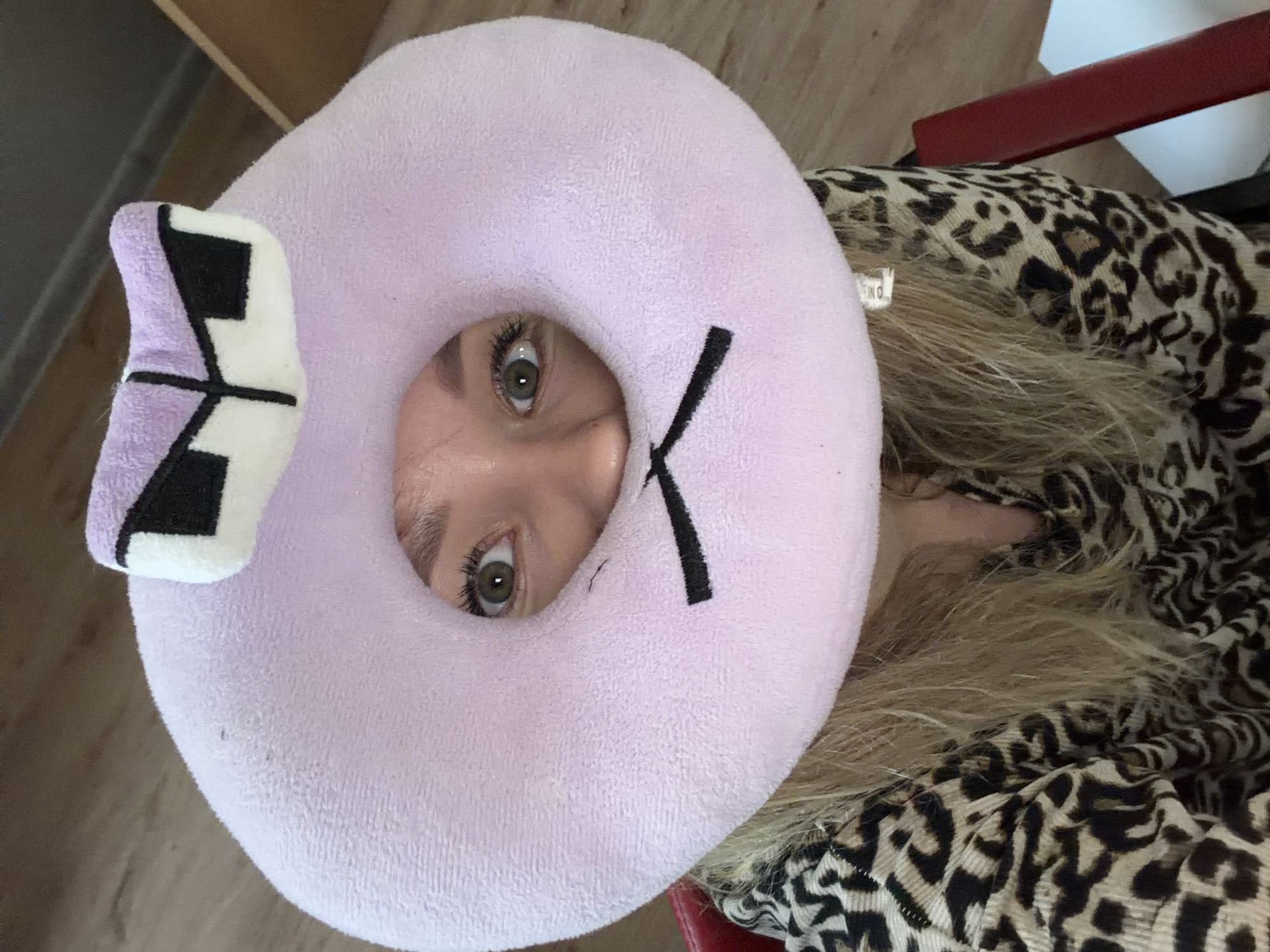 A light-skinned person is looking up through the central opening of a lavender-colored, circular pillow, which has a small, stylized, purple and black ear on one side and a black, angled line marking on the opposite side. The person has light brown eyes and blond hair. The person is wearing a brown and black animal print top. The pillow is on a red-painted chair in a room with brown flooring and a white wall. There is no evidence in the image to support that the location is Chaikovsky, Russia.