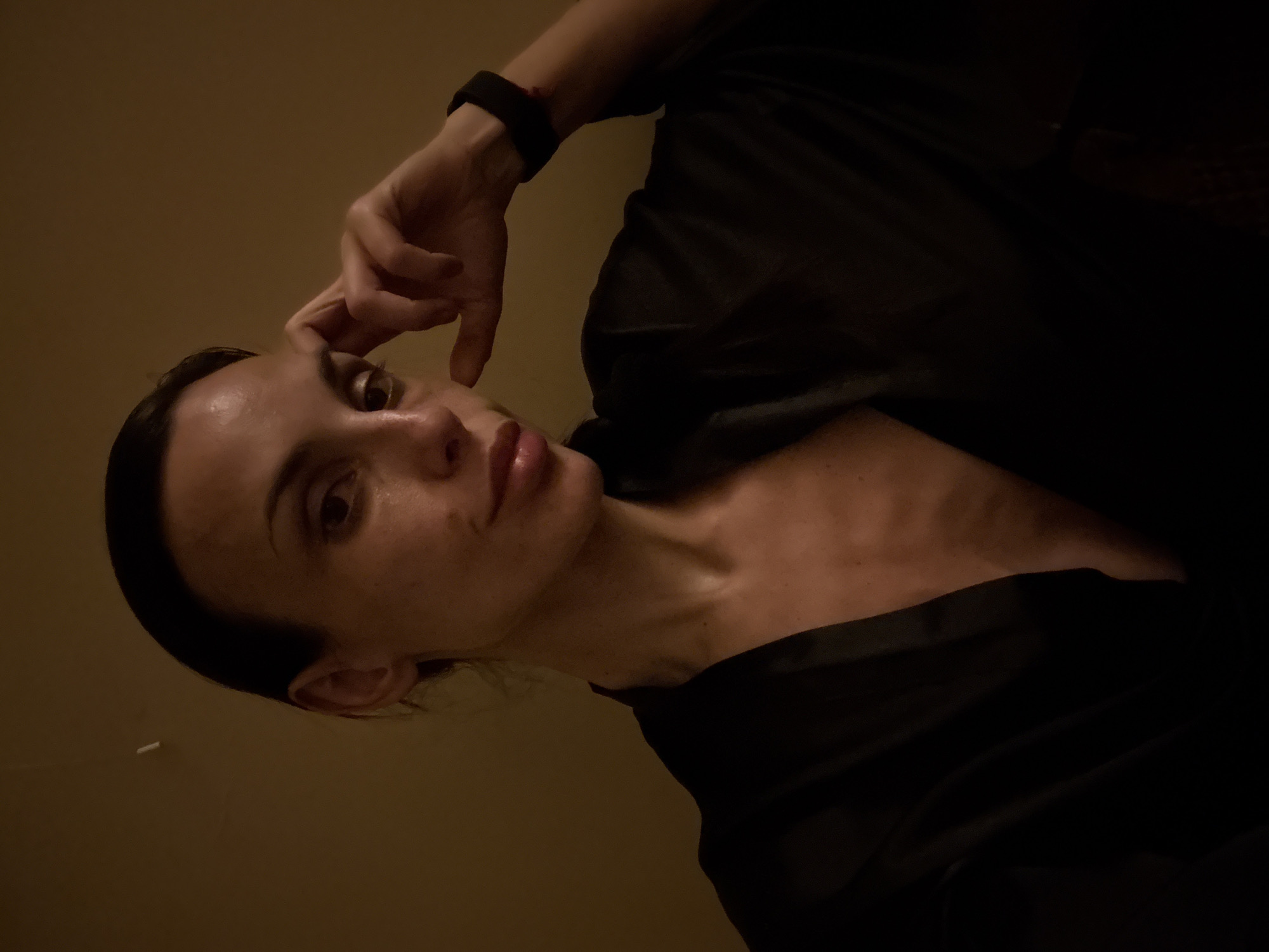 A woman with dark hair is depicted from a low angle, wearing a dark, unbuttoned robe or garment that reveals part of her chest and collarbone. Her right hand is raised to her forehead, appearing to be in a pensive or contemplative pose. She is wearing a black wristband. The background is a plain, dark beige or tan. There is no information in the image to indicate that it was taken in Pleven, Bulgaria.