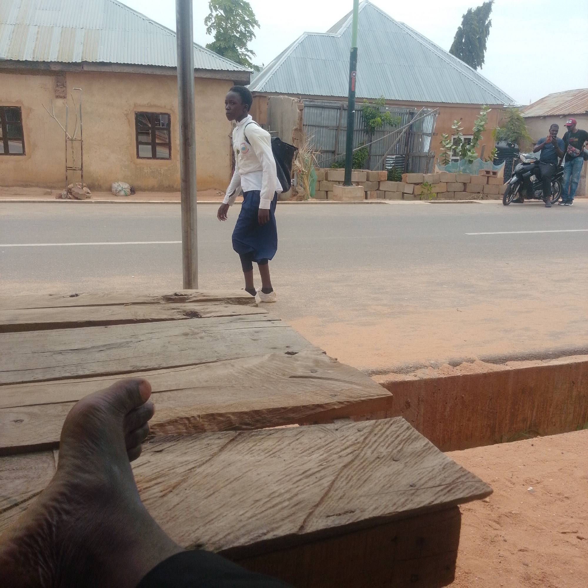 The image shows a young person walking along a dusty roadside in what appears to be a rural or semi-urban area of Gombe, Nigeria. The person, dressed in a white collared shirt and a dark blue skirt over leggings, carries a black bag over their shoulder. They are walking past a series of buildings with corrugated metal roofs and stucco walls. The road has a white line marking the center.
In the background, two men are standing near a motorcycle. One man, wearing a blue shirt, is looking at his phone, while the other, in a black shirt and red cap, is looking towards the camera. There are stacks of cinder blocks and some greenery near the buildings. The foreground features a close-up of a person's feet resting on a wooden surface, suggesting the photo was taken from a seated position. The ground is a mix of sand and dirt. The overall scene suggests a bright, possibly warm day. The environment appears to be in Nigeria, given the location context. No visible text is present in the image.