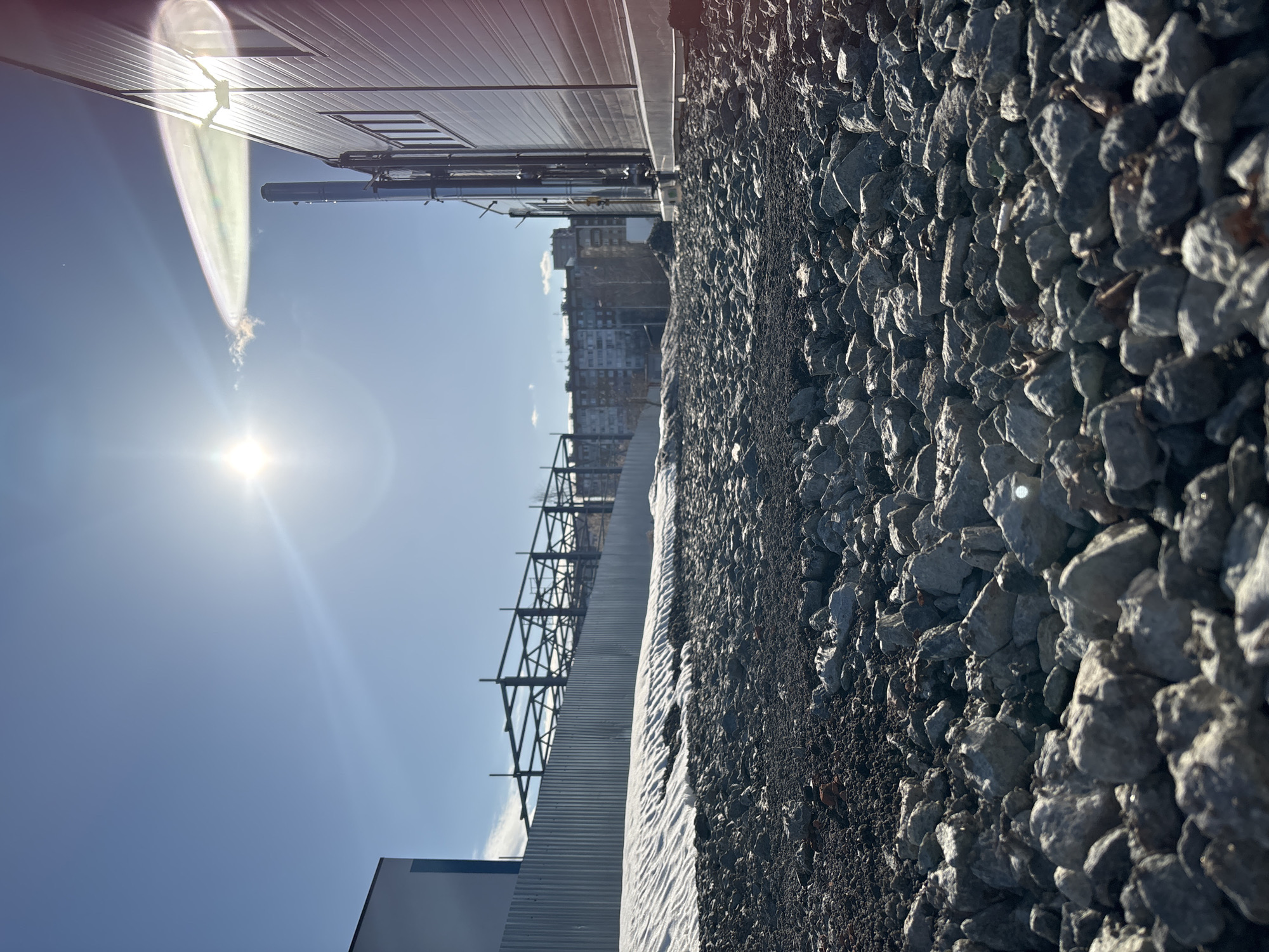 The image shows an outdoor scene under a bright sun. The foreground is dominated by a bed of grey rocks or gravel. Adjacent to the rocks is a lower corrugated metal structure, followed by a metal frame construction. In the background, blurred, are multi-story buildings. The architectural style appears modern. The photograph is taken at a low angle. Based on the architectural style and the presence of modern construction, the location is likely Izhevsk, Russia. No people are visible.