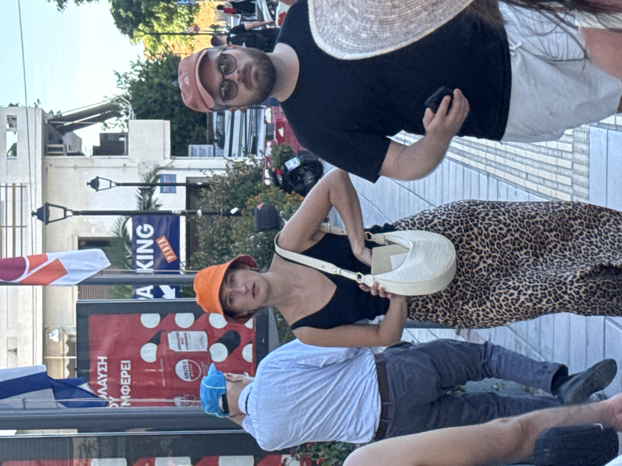 The image captures a street scene in Chania, Greece, focusing on a group of people walking along a pedestrian walkway. In the foreground, a woman wearing an orange bucket hat and a leopard print skirt stands with her body turned slightly to the right. She is holding a white, textured handbag on her shoulder. To her right, a man with a beard, wearing a black t-shirt and a reddish baseball cap, is walking forward. He is holding a dark object, possibly a phone, in his right hand. Behind the woman, a man in a light blue shirt and a blue cap is walking away from the camera. Various flags and signage are visible in the background, including a Greek flag and advertisements. The environment appears to be a commercial area with buildings and streetlights. The walkway is paved with light-colored tiles.