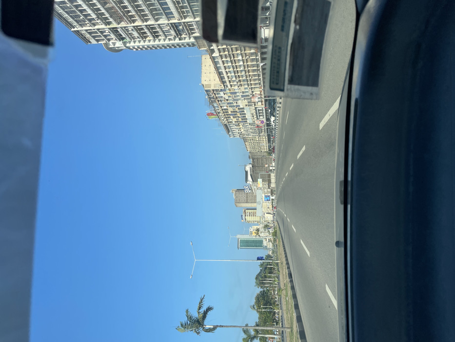 The image presents a sun-drenched urban street scene in Luanda, Angola, captured from inside a vehicle looking forward and slightly to the left. The sky is a vast, cloudless blue, indicating bright daylight, likely midday or early afternoon.
On the left side of the frame, a continuous stretch of multi-story buildings lines the street, receding into the distance. The closest structure on the far left is a very tall, modern building with numerous windows or balconies. Further along, other buildings, some appearing to be under construction or featuring extensive external structures, are visible. A small, colorful flag with red, green, and black stripes can be seen on one of these more distant buildings, possibly the Angolan flag. The word "BANK" is clearly visible on a building further down the street, and letters resembling "ANB" or "AMC" are discernible on another.
The right side of the image is dominated by a wide, paved road with multiple lanes delineated by solid and dashed white lines. The road extends straight into the horizon. A median strip, featuring sparse green vegetation and streetlights, separates the road from the buildings or perhaps a parallel carriageway. Several palm trees are scattered along this median and further down the roadside, contributing to a tropical aesthetic.
In the immediate foreground on the far right, a dark gray or black section of the vehicle's interior, likely a door panel or dashboard, partially frames the scene. A small, rectangular white object, possibly a sticker or card, is attached to this interior part, bearing faint, illegible text. The overall scene depicts a modern cityscape under clear and sunny weather conditions.