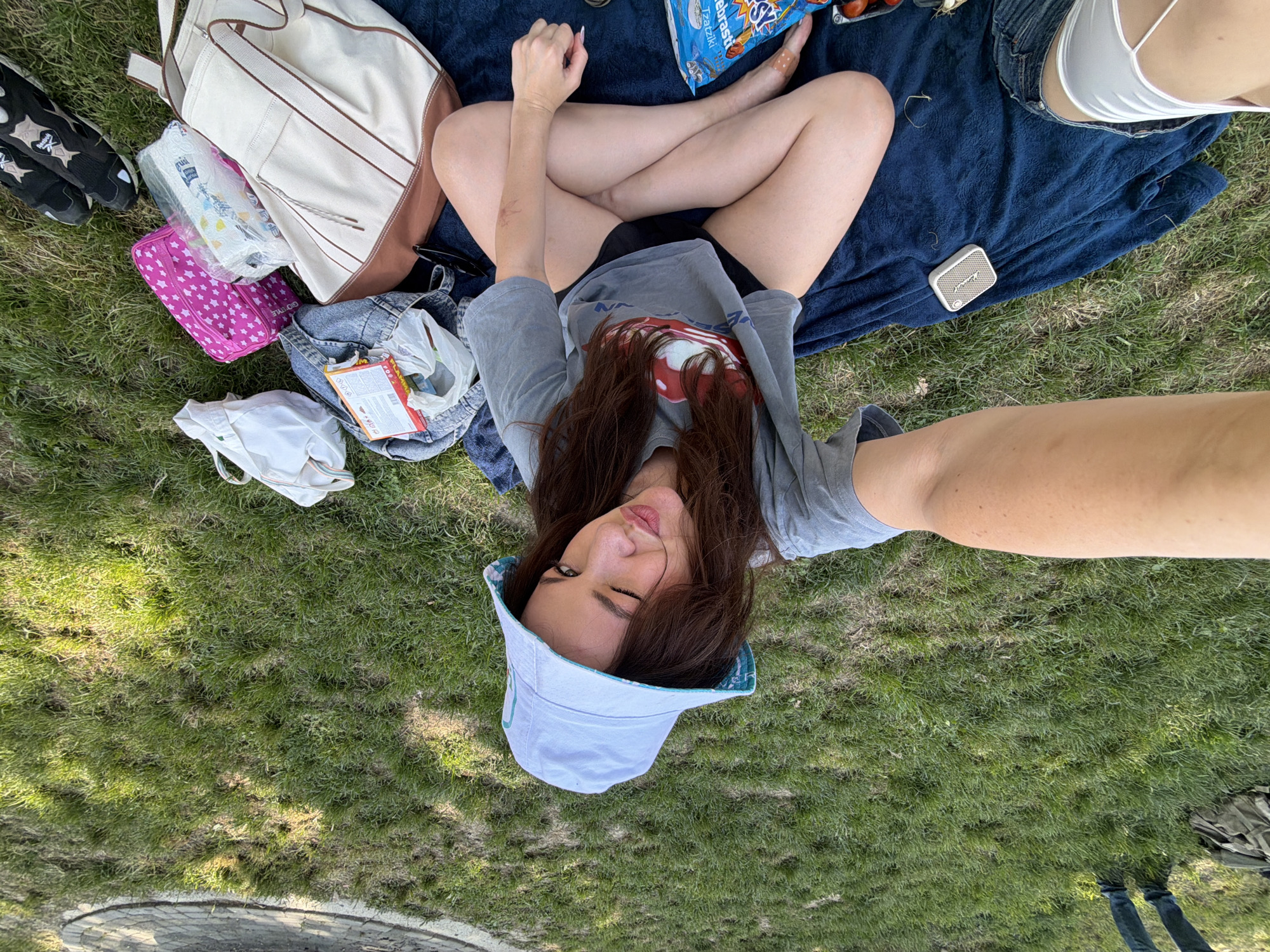 A young woman wearing a light blue bucket hat and a gray T-shirt is lying on a blue blanket in a grassy area. The blanket has various items scattered around it, including a white backpack, a pink bag, a bag of potato chips, a pair of black shoes, and a denim jacket. The woman is positioned in the center of the image, taking a selfie with her arm outstretched. The location appears to be a park or green space within the Cukarica Urban Municipality, Serbia, evidenced by the natural setting of grass. The overall impression is a casual, leisurely scene of relaxation in a park.