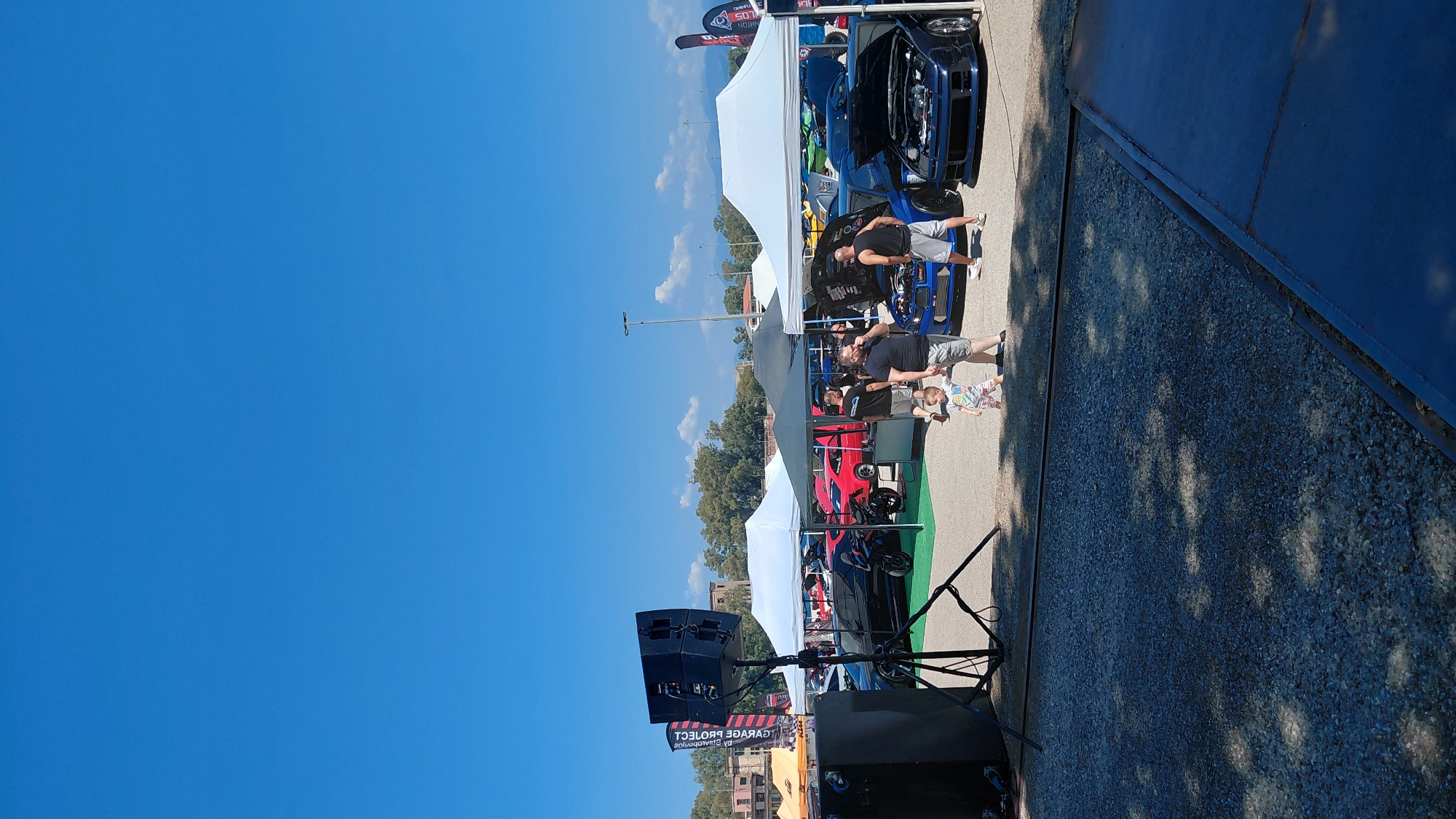 This vertical, eye-level outdoor photograph captures a sunny day at what appears to be a car show or exhibition, likely in the Municipal Unit of Aegio, Greece. The clear blue sky occupies the upper portion of the image. In the foreground, a gravel-covered area is visible, with shadows cast by trees creating dappled patterns. To the left, a large black speaker system is mounted on a tripod.
Several tents are set up, providing shade for displays. Underneath a white tent, a black motorcycle and a red car are parked. Another black tent is also visible. In the mid-ground, a blue car with its hood open is on display, with a man and a child standing nearby. Further to the right, another blue car with its hood open is also showcased. Several other cars, their models indistinct, are present further back under tents. A few individuals are scattered around the area, some interacting with the cars. The background reveals trees and some buildings, suggesting an urban or semi-urban setting. Two tall flagpoles with banners are visible on the left. The overall scene depicts a public event with vehicles and attendees.