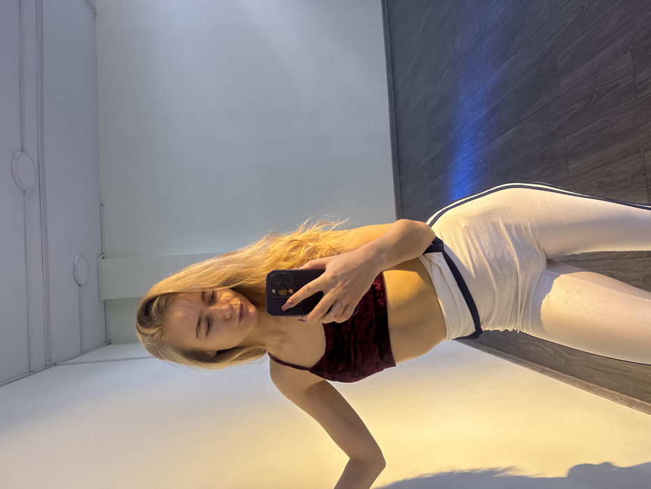 The subject, a young Caucasian female, is depicted indoors within what appears to be a dance studio or gymnasium, location Unknown, Unknown. She has long, wavy blonde hair and is wearing a dark red velvet sports bra and light-colored athletic leggings with dark vertical stripes along the sides and a contrasting waistband. Her right hand holds a dark-colored smartphone, positioned to capture a selfie; her fingernails are painted a dark shade. Her left arm is bent, with her hand resting against a plain white wall, which is illuminated by warm, golden light. The environment includes a dark wood-like floor and a white ceiling fitted with circular light fixtures. A large mirror occupies the right side of the image, reflecting the room. A faint blue light streak is visible on the floor towards the right.
