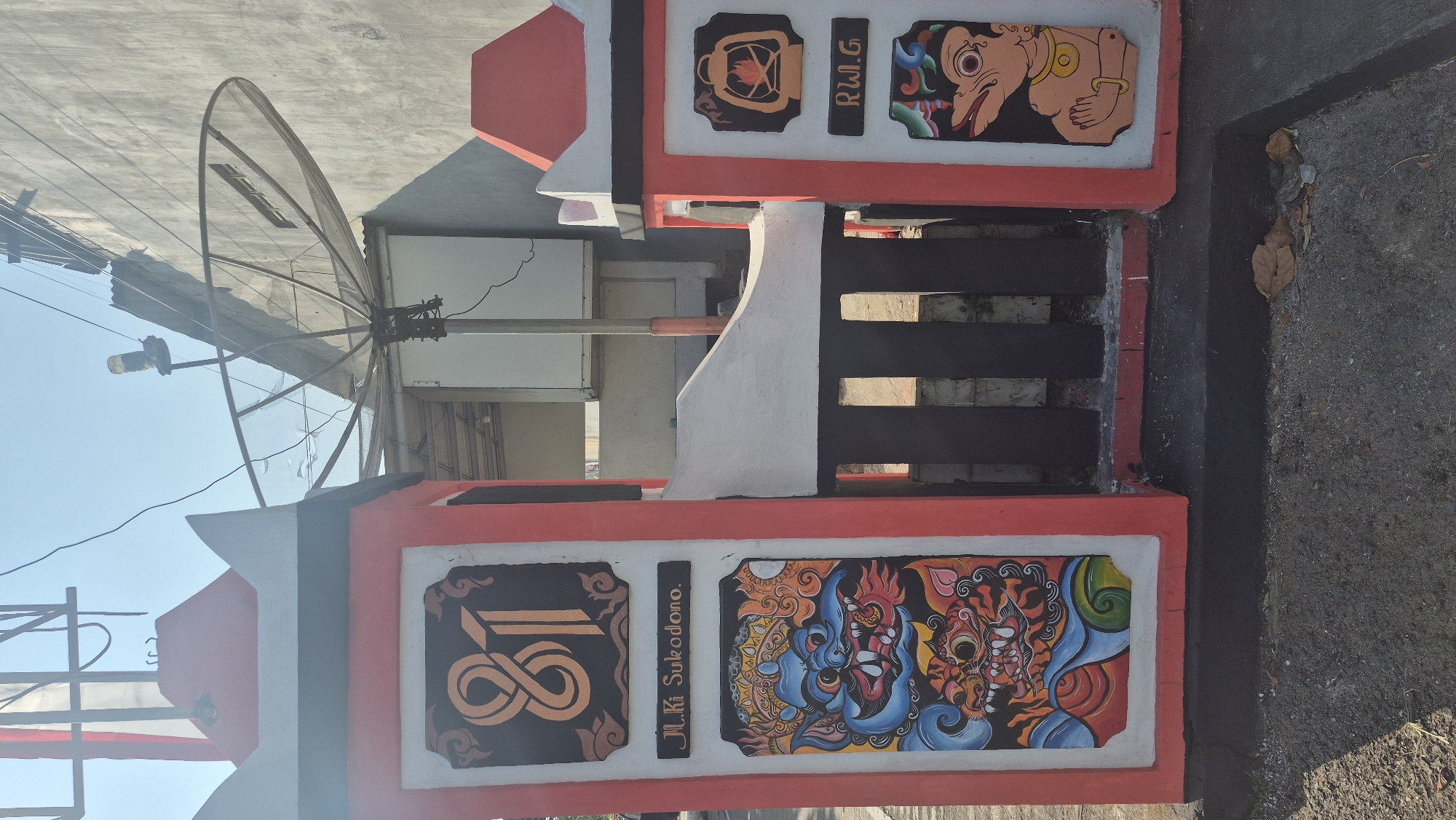 A daytime, eye-level, medium shot captures a gate entrance adorned with vibrant traditional Indonesian artwork. The gate is constructed from concrete pillars painted red and white, with a black, barred entrance. The left pillar features a large numeral "81" in orange, followed by the text "Jl. Ki Sukodono" in black. Below this, a colorful mural depicts two mythical creatures, one with blue and red scales and sharp teeth, and another with orange and black stripes. The right pillar has an illustration of a lantern with a flame, followed by the text "R.W.G." and a depiction of a puppet character with a light brown face and yellow adornments. The background reveals a grey building, a satellite dish, and a clear blue sky. The ground in front of the gate is paved with asphalt and scattered with dry leaves. The scene is located in the city of Limbangan, Indonesia.
