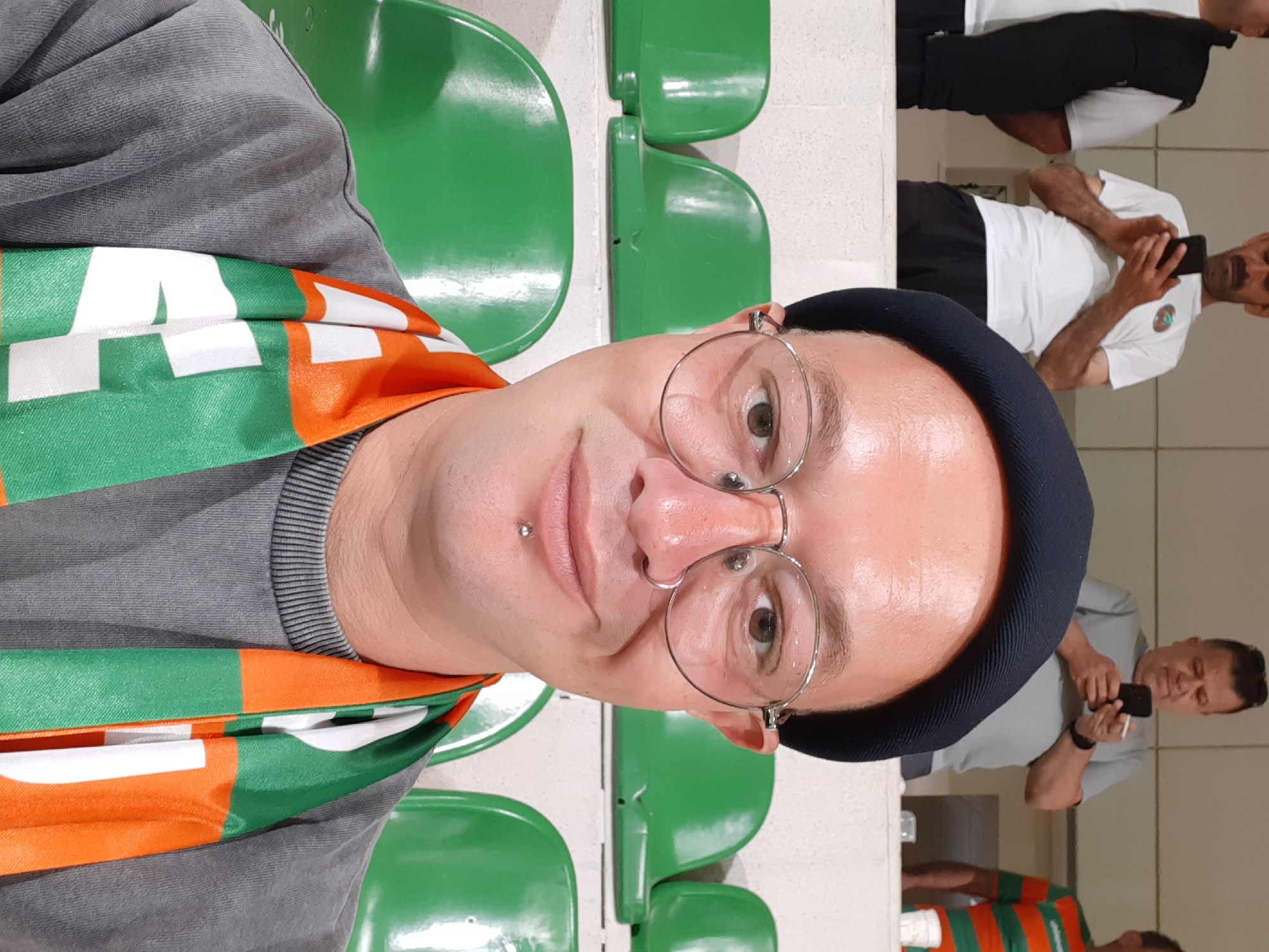 A medium close-up shot depicts a person wearing round glasses, a black beanie, and an orange, white, and green scarf, possibly a sports team scarf. The person has a lip piercing and is seated in a green plastic seat, likely in a stadium or arena. In the background, other individuals are present, some wearing white shirts and dark pants, some using mobile phones. Based on the scarf colors (orange, white, and green), this is likely the Alanyaspor team. Given Alanyaspor's location, it is highly probable that this photo was taken in Alanya, Turkey. The background is slightly blurred, suggesting the camera is focused on the person in the foreground.