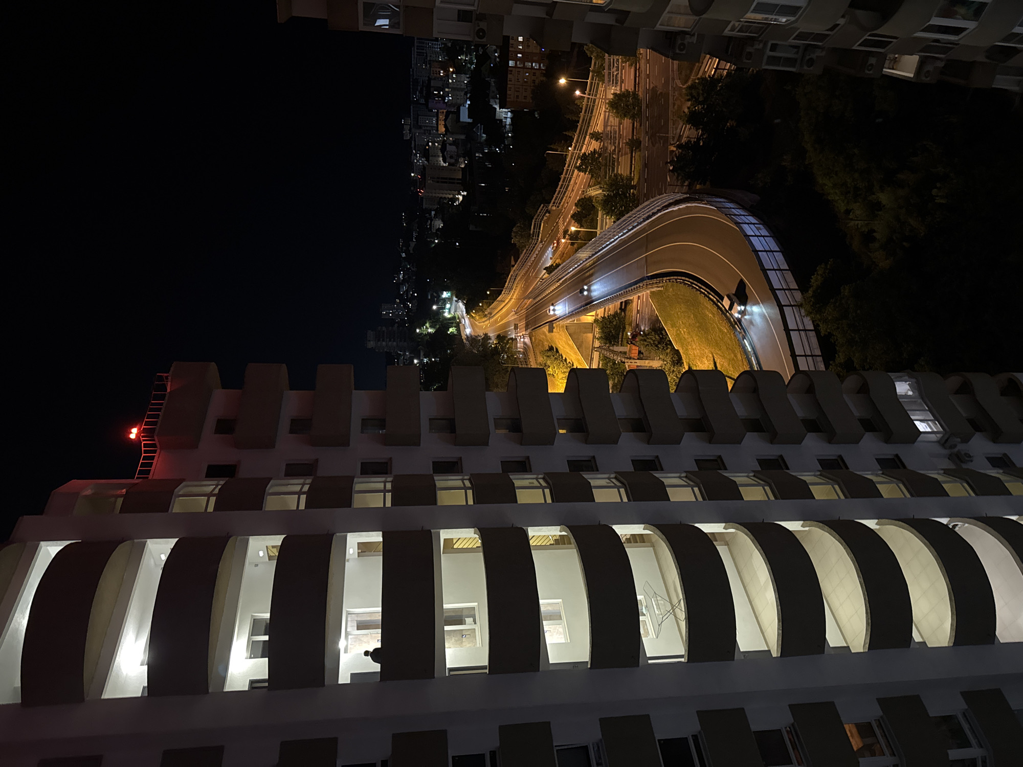 The image is a nighttime aerial view of a city, likely in Sochi, Russia, featuring a multilane highway curving through a landscape of buildings and trees. On the left, a section of a modern, white apartment building with curved balconies is prominently visible, its balconies illuminated from within. One balcony on the upper floors shows a silhouetted human figure standing near a doorway. The building on the right is partially visible and appears to be similar in design. The highway below is well-lit by streetlights, and several cars are visible, their headlights and taillights creating streaks of light on the wet road surface, suggesting recent rain. The overall environment is urban with lush greenery interspersed between structures. The sky is dark, indicating nighttime.
