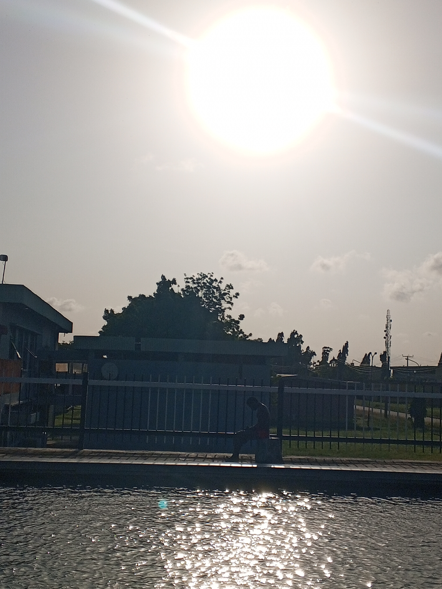 A sun-drenched, semi-obscured scene featuring water in the foreground with intense sunlight reflecting off its surface. In the midground, there's a fence and behind it, a building and trees. A person is seated on a ledge near the fence. In the background, communication towers and power lines are visible. The location is possibly Tawpo, Nigeria, based on the prompt's details.