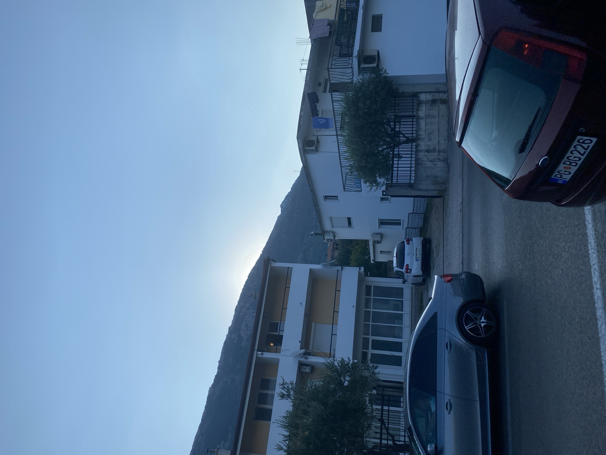 The image depicts a street scene in Sutomore, Montenegro, at what appears to be dawn or dusk, given the low angle of the sun peeking from behind a mountain range in the background. Two residential buildings are visible, one to the left and one to the right of the frame, with a road running in front of them.
The building on the left is multi-storied, featuring a pale yellow facade with white balconies and window frames. A mature tree with dense foliage stands in front of it. The building on the right is also multi-storied and painted white. It has balconies with railings, and some laundry is seen drying on one of the balconies. Both buildings appear to be typical of a coastal town.
Several cars are parked on the street. In the foreground, a dark red hatchback is partially visible on the right, showing its rear end with a license plate reading "PG BG 226". To the left of this, a silver sedan is seen from its side profile, with its wheels and lower body clearly visible. Further down the street, a white hatchback is parked in front of the building on the left, displaying a license plate "BR DU147".
The sky is a pale blue, indicating either early morning or late evening light. The overall impression is one of a quiet street in a town situated near a mountainous landscape. The road appears to be paved and wet, possibly from recent rain or dew.