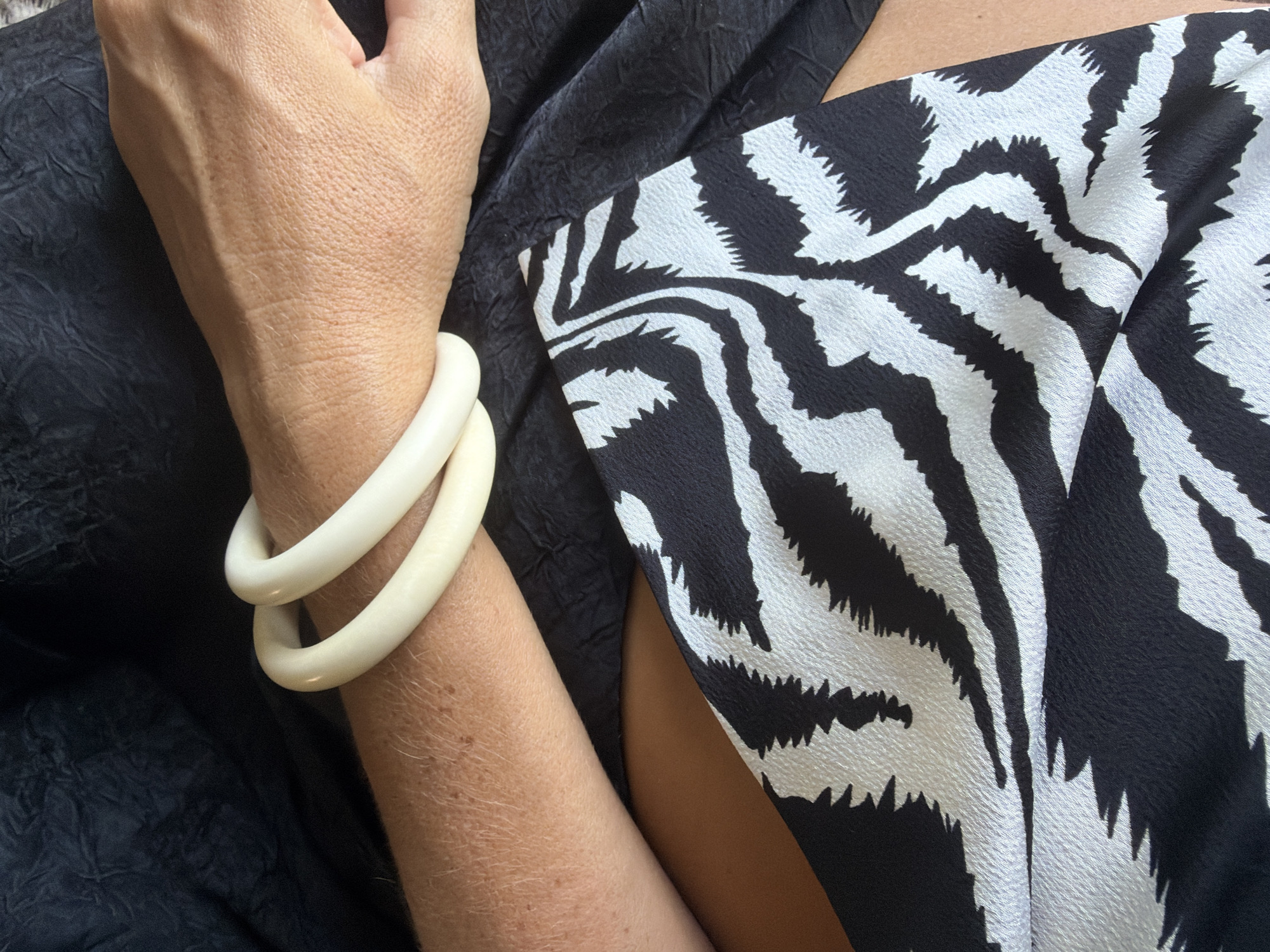 An overhead, close-up shot shows a person's arm and hand adorned with two off-white, resin bangles. The arm and hand are resting on a textured black fabric. Below the arm, a black and white zebra-print fabric is visible, folded and creased. The scene is lit by natural light, creating subtle shadows and highlighting the textures of the fabrics and skin. The location in the city of Ko Samui District, Thailand, is not discernable from the provided image.