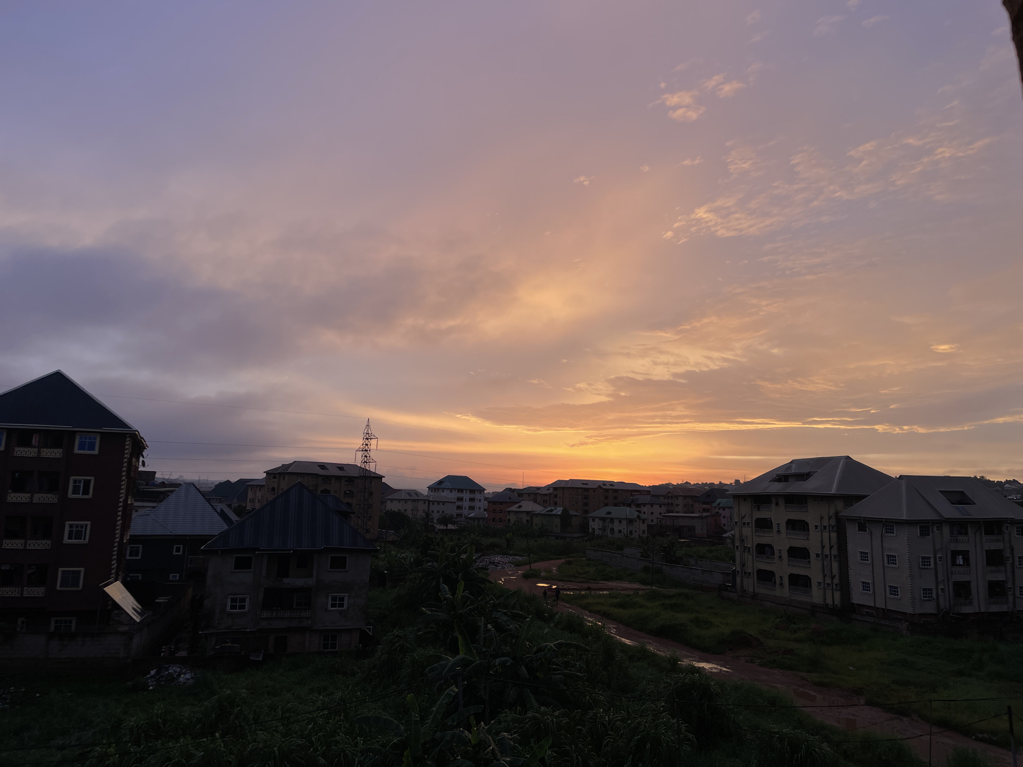 The image captures a cityscape at dusk in Nkpor, Nigeria. The sky is a blend of purples and oranges, with clouds illuminated by the setting sun. In the foreground, dense green foliage, including what appear to be banana plants, obscures the lower portion of the scene. Several multi-story buildings are visible, featuring various architectural styles and colors, some appearing aged and others more modern. One prominent building on the left is dark red with visible windows and balconies. To its right, a darker building with a sloped metal roof is partially visible. In the middle ground, a dirt road winds through patches of green grass, with puddles of water indicating recent rain. Two small figures can be seen walking on this road. Further back, more buildings of varying heights and designs are scattered across the landscape. A tall electricity pylon with wires is also present in the background, a common sight in urban Nigerian settings. The overall impression is of a residential area experiencing the transition from day to night.