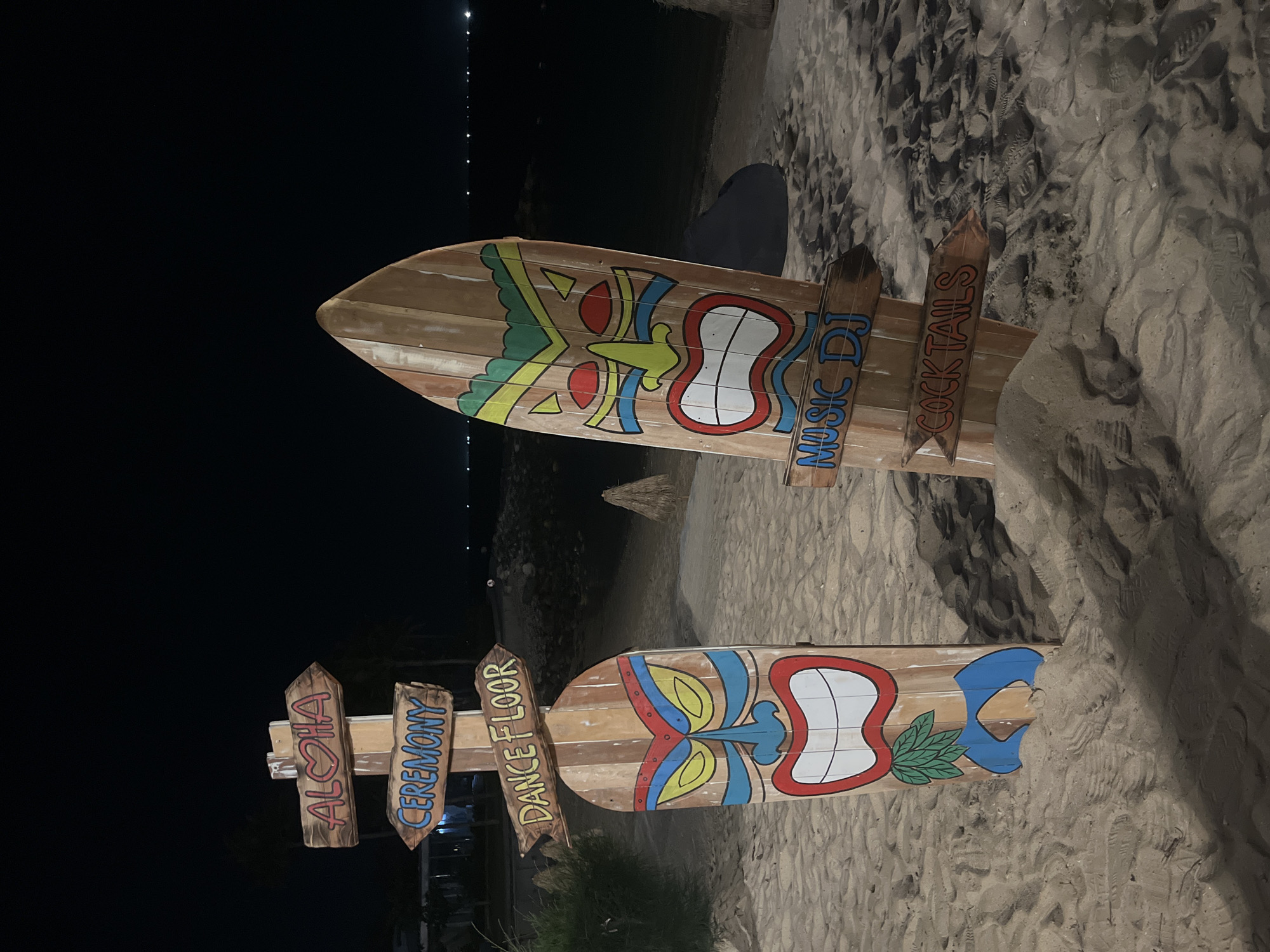 This image depicts a beach scene at night in Nha Trang, Vietnam. Two wooden surfboards painted with stylized tiki faces are planted in the sand. The surfboard on the left has three wooden directional signs attached, reading "ALOHA" with a heart symbol, "CEREMONY," and "DANCE FLOOR." The surfboard on the right has two wooden directional signs, reading "MUSIC DJ" and "COCKTAILS." The sand is disturbed with footprints and there are faint lights visible in the background, suggesting the presence of buildings or a town along the coast. The sky is dark, indicating nighttime.