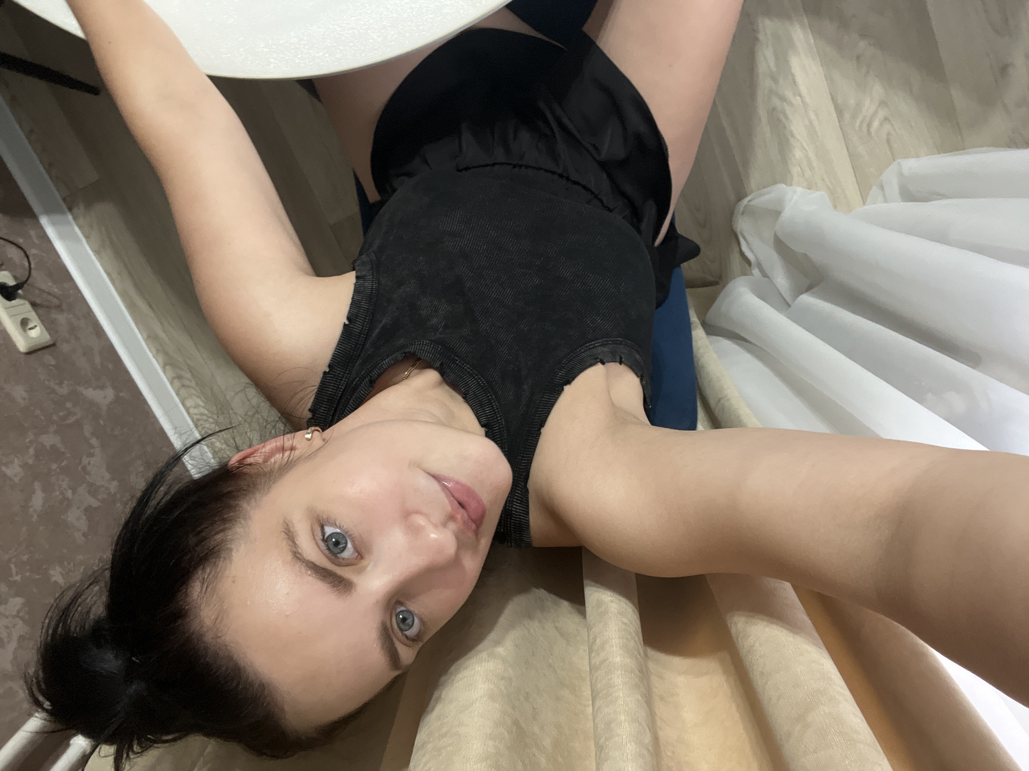 A young woman with dark hair pulled into a bun and striking blue eyes is lying on her back, looking up at the camera. She is wearing a black tank top and dark shorts. Her right arm is extended upwards, resting on a white table. The scene appears to be indoors, with a light-colored wooden floor and beige curtains visible in the background. A white electrical outlet is mounted on the wall to the left. The lighting suggests it is daytime. The woman's expression is relaxed and her gaze is direct, with a slight smile. The image is taken from a high-angle perspective. No visible text is present.