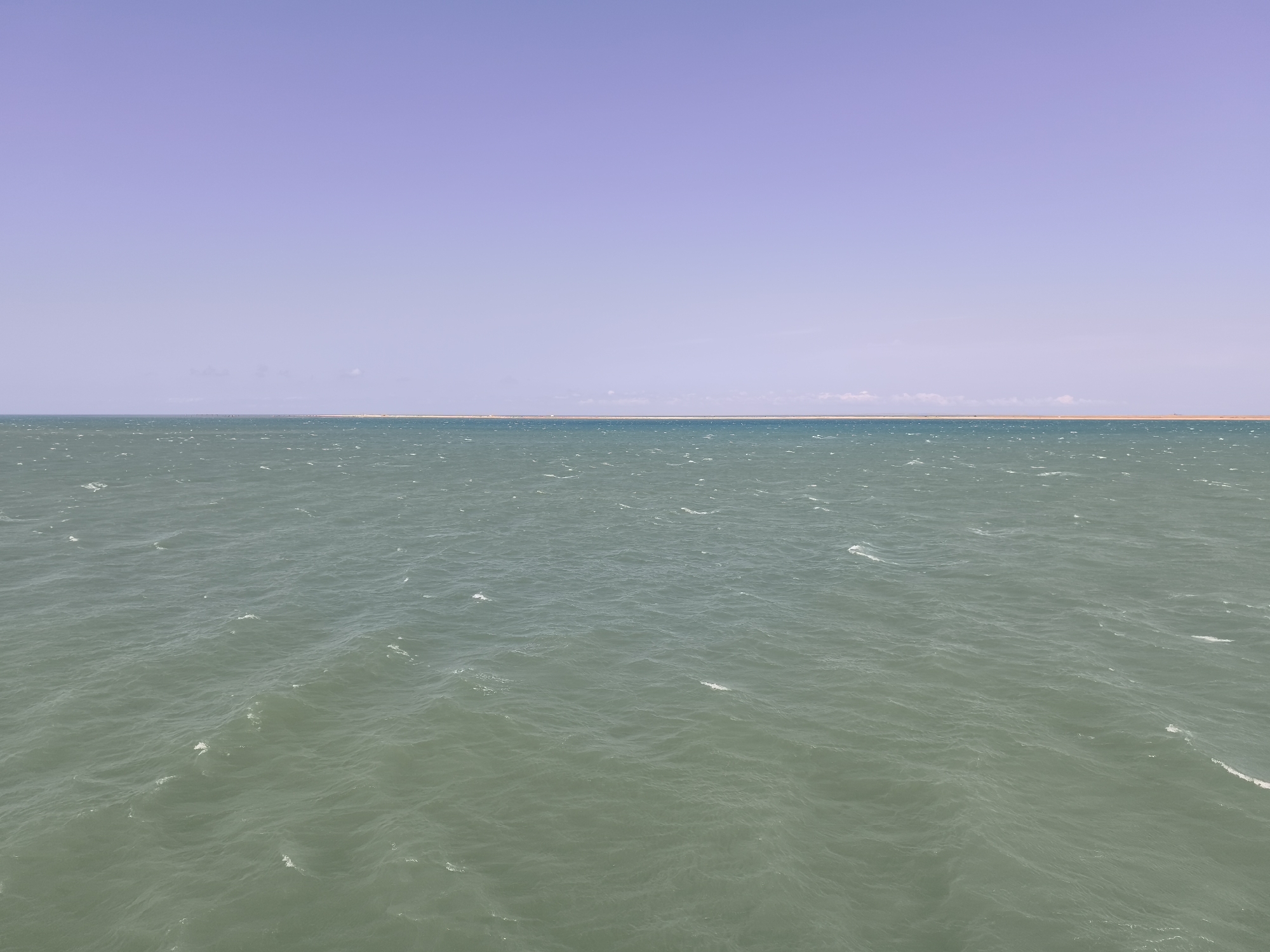 The image depicts a panoramic view of the Caspian Sea from Pirallahi Raion, Azerbaijan. The foreground is dominated by the choppy, greenish-blue waters of the sea, with small whitecaps indicating a moderate breeze. In the midground, a narrow strip of land, likely the coast of Pirallahi Island or a nearby mainland area, is visible as a sandy, light-colored line. Beyond the landmass, a faint, hazy horizon suggests a distant coastline or further expanse of the sea. The sky above is a clear, pale lavender, with no clouds visible. The overall impression is one of a wide, open seascape under a serene sky. There are no people, animals, or man-made structures clearly identifiable in the scene, emphasizing the natural environment.