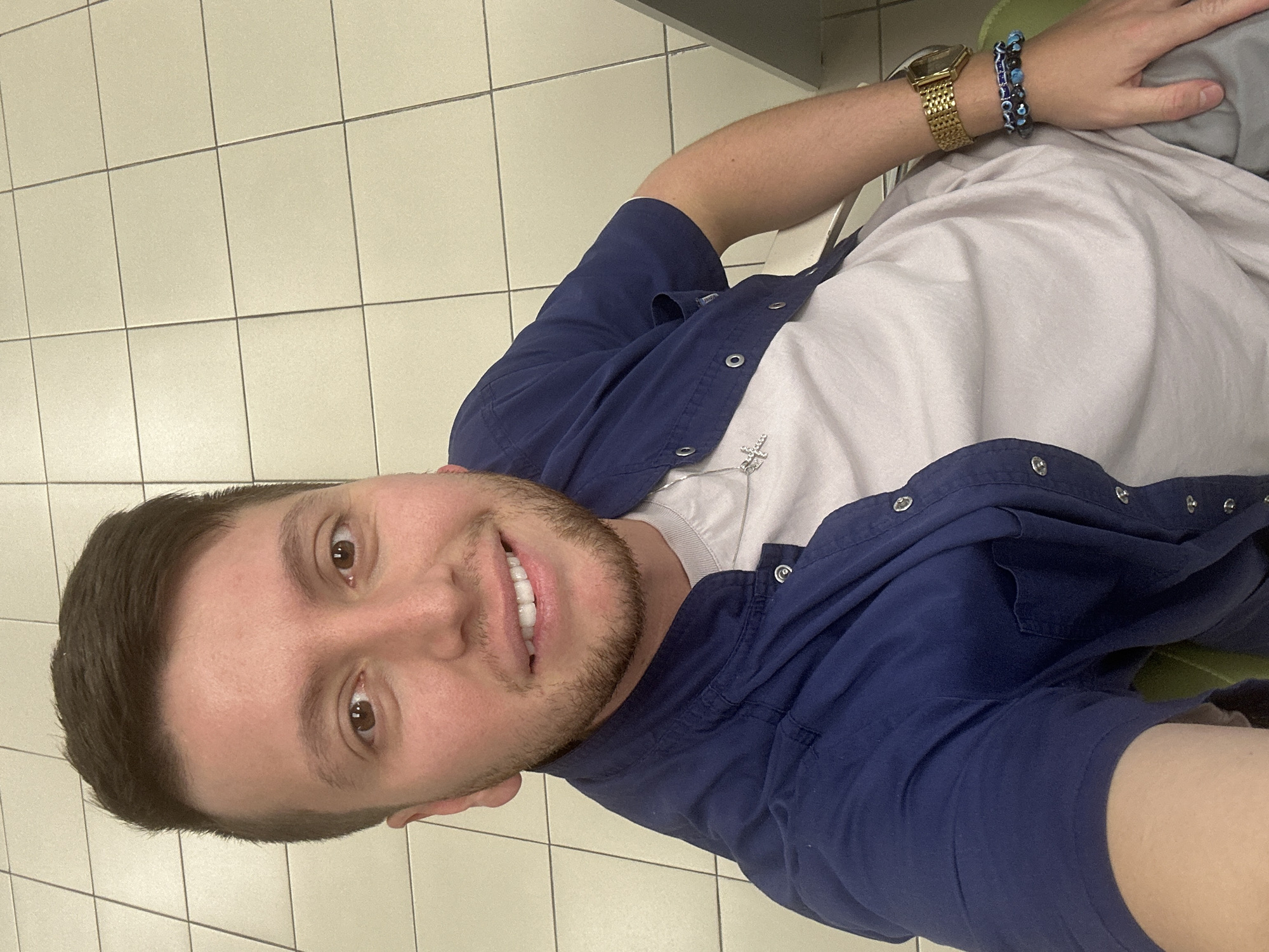 A young adult male is captured in a selfie taken indoors, in what is identified as Moscow, Russia. The man is positioned diagonally in the frame, looking directly at the camera with a wide, cheerful smile that reveals his white teeth. He has short, neatly styled brown hair, brown eyes, and light stubble on his face.
He is dressed in a dark blue short-sleeved button-up shirt, worn unbuttoned over a light gray or white t-shirt. A silver-colored necklace with a small cross pendant is visible against his t-shirt. On his left wrist, he sports a gold-colored digital watch alongside two beaded bracelets, one featuring blue "evil eye" beads. A segment of dark blue shorts is visible towards the bottom right of the image.
The setting appears to be an indoor space with light-colored square tiles and dark grout lines covering the floor. A dark green or gray surface, possibly a piece of furniture like a chair or bench, is partially visible in the background. The lighting suggests an artificial indoor environment. The man's expression conveys happiness and relaxation. There is no visible text in the image.