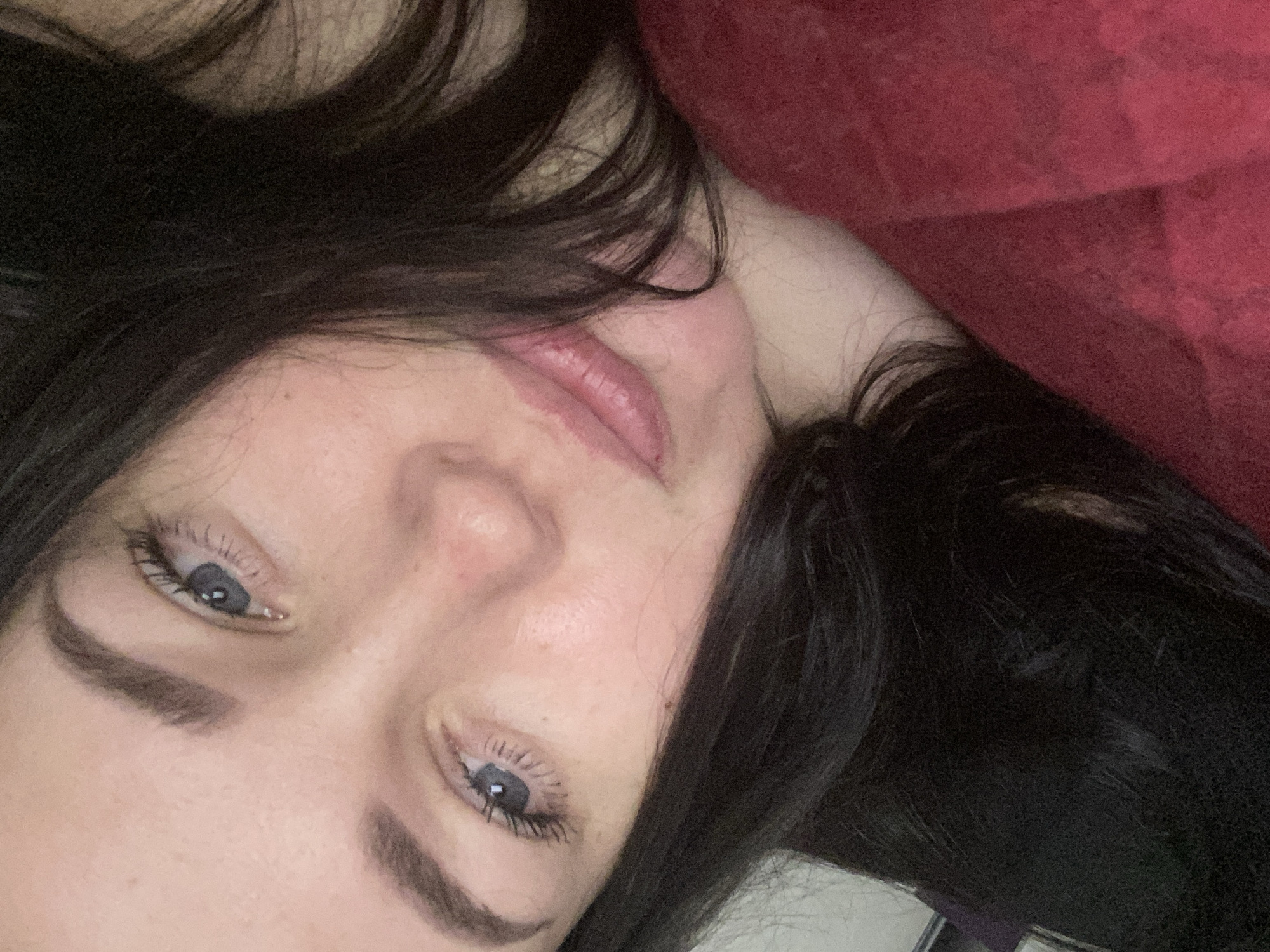 The media file displays a close-up, angled view of a person, likely female, with dark hair. The subject is seen from the upper chest and head, lying down, with their head tilted. Their light-colored eyes, possibly blue or grey, are open and directed towards the viewer. Long, dark eyelashes are visible, along with well-defined eyebrows. The skin tone is fair.
The person's lips are slightly parted, revealing a natural pink hue. Dark, wavy hair frames the face and spills around the head. A section of a red, textured fabric, resembling a blanket or warm garment, is visible in the upper right portion of the image, suggesting an indoor and comfortable setting. No specific activity beyond posing for the camera or resting is discernible. The overall impression is one of a relaxed, contemplative pose. There is no visible text in the image.