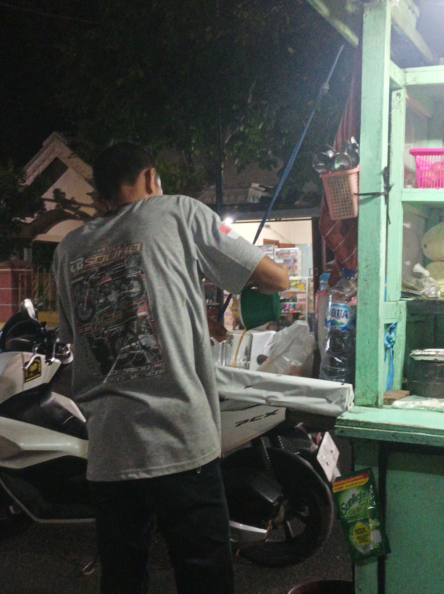 The image depicts a street scene at night in Nganjuk, Indonesia, likely near a small roadside food or drink stall. A person is standing beside a motorcycle, their back to the camera, wearing a grey t-shirt with a graphic print featuring motorcycles. The person appears to be interacting with the stall, which is characterized by a green wooden frame. On the stall's shelves, various items are visible, including a pink basket, a green bottle of "AQUA" mineral water, and a pouch of "Sunlight" dish soap. The background shows a building with a peaked roof and trees, illuminated by ambient light. The ground is paved and appears to be a street or walkway.