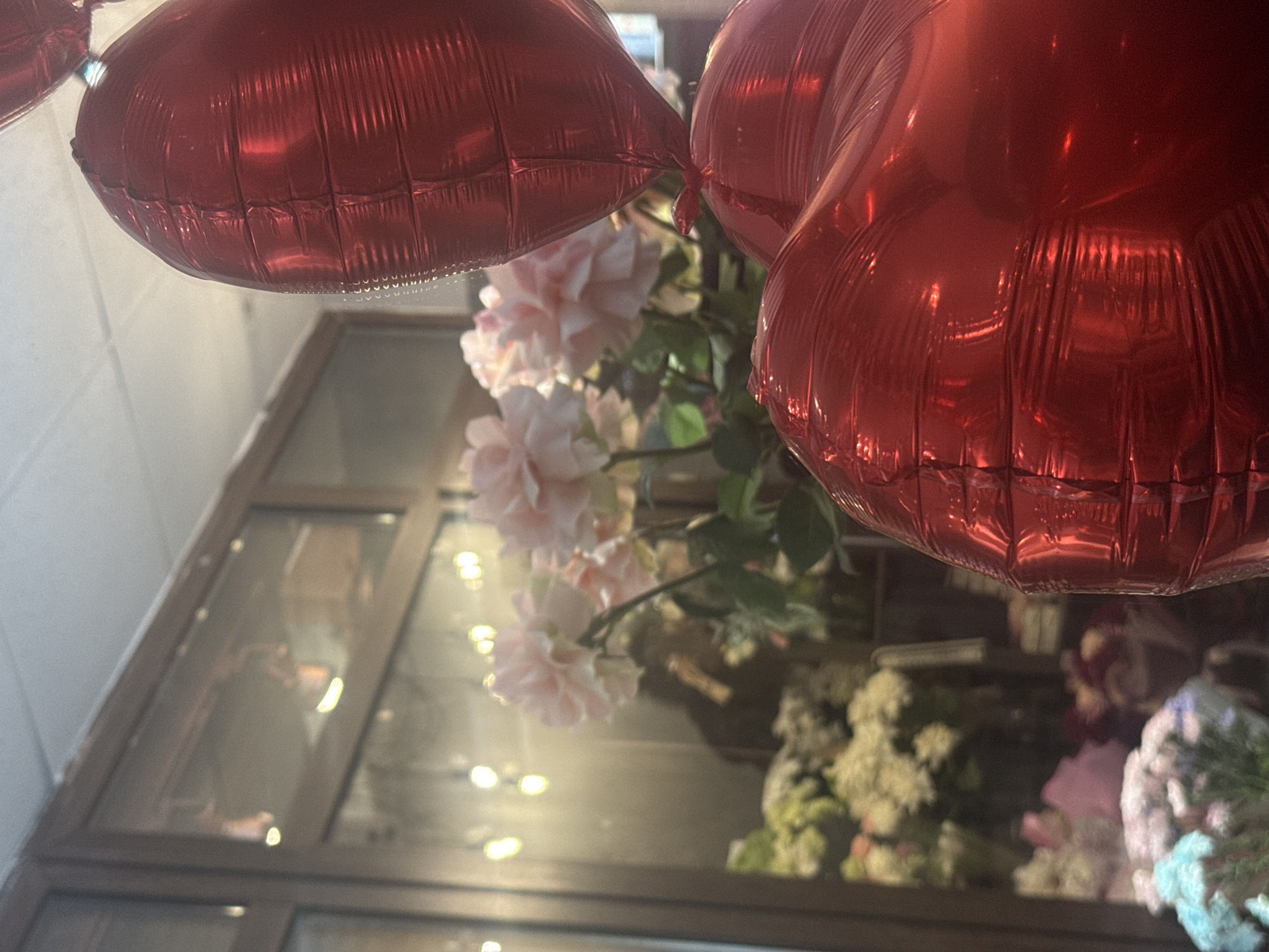 The image captures an indoor scene with two inflated, red heart-shaped balloons dominating the upper portion of the frame. Behind the balloons, a bouquet of light pink flowers with green foliage is visible. The scene is reflected in a glass-paneled window or display case, which also shows reflections of people inside the space, possibly a store or business. Other floral arrangements of varying colors and types are visible through the glass, suggesting the location could be a florist shop. There is no definitive evidence to confirm the exact location within the city of Perm, Russia.
