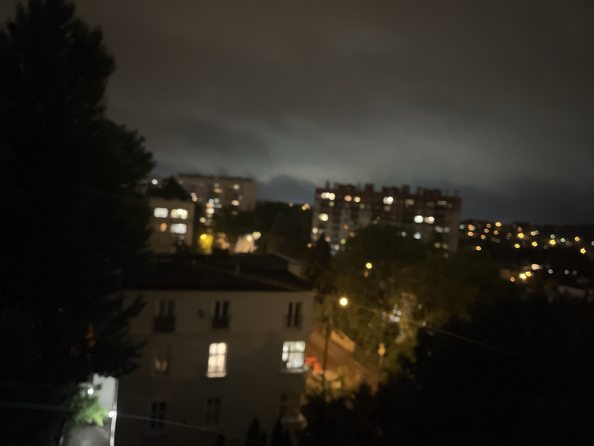 The media file presents a blurry, high-angle nighttime view of an urban or suburban landscape. A large, dark tree silhouette occupies the left side of the foreground, partially obscuring the scene.
In the midground, several multi-story buildings are visible; one prominent lighter-toned building in the lower-middle features illuminated rectangular windows, while other larger, darker blocks further back on both left and right also display numerous lit windows. Distant streetlights and building lights create soft, circular bokeh patterns across the background, indicating a vast expanse of city lights.
The sky above is dark and heavily overcast, with indistinct cloud formations discernible, likely illuminated from below by city glow. No specific activities or people are depicted, giving the scene a static and tranquil quality. The overall impression is one of quiet urban night, viewed from an elevated perspective such as a window or balcony. There is no visible text.