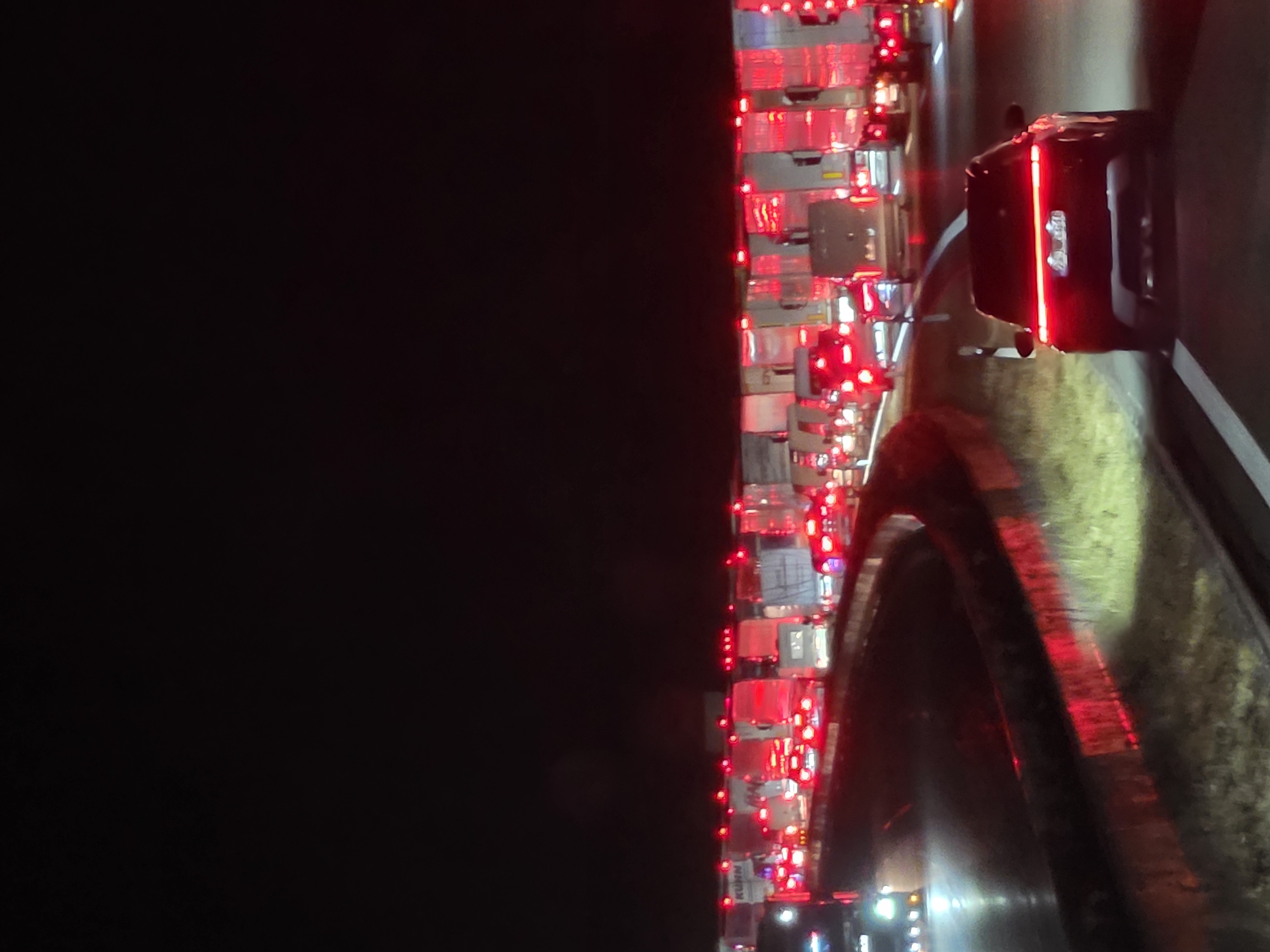 The image depicts a nighttime traffic jam on a highway, likely the A3, near Uettingen, Germany. A dark-colored SUV with illuminated taillights is in the foreground, facing away from the viewer and traveling in the right lane. Behind the SUV, numerous trucks are lined up, their red taillights creating a vibrant glow against the dark asphalt. The highway appears to be multi-lane, with vehicles moving slowly or stopped. The environment is characterized by the darkness of night, with the only significant light sources being the vehicle lights and some roadside illumination. The scene conveys a sense of congestion and standstill traffic.