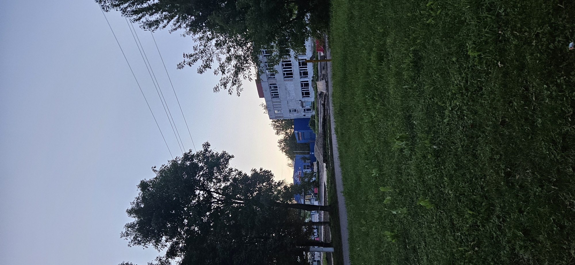 The image depicts a view of an industrial or commercial area in Vitsebsk, Belarus, at dusk. In the foreground, a patch of green grass is visible. Beyond the grass, there is a paved pathway or road. In the middle ground, several buildings are present. One building on the right is a multi-story structure with many windows and a light-colored facade. To the left of this building, there are two bright blue, low-rise structures. Between these buildings, there are trees and a pile of dirt or sand, suggesting ongoing construction or maintenance work. Power lines are visible across the sky. The sky is a pale blue, indicating the end of the day. There are no people or vehicles clearly visible in the scene.