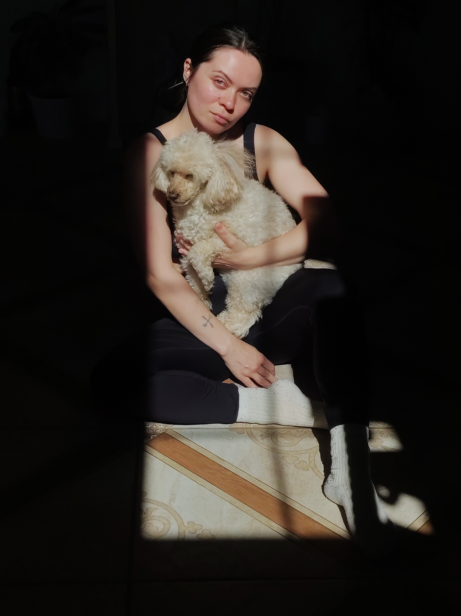 A young woman sits cross-legged, holding a small, white poodle dog in her arms. She is wearing a black tank top, dark leggings, and white socks. A tattoo is visible on her left forearm. The scene is sharply divided by light and shadow, with a tiled floor visible in the sunlit area. The location is Фоки, Russia.