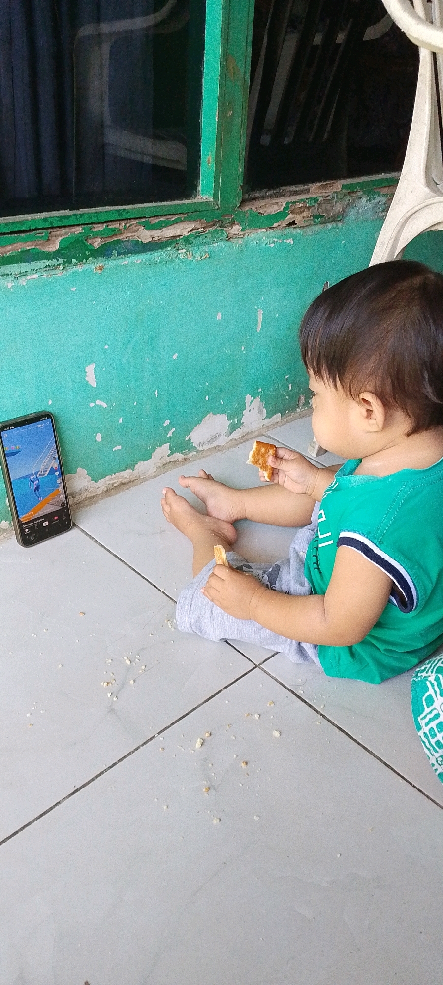 An infant is sitting on a tiled floor next to a teal-colored wall with peeling paint. The infant is wearing a green t-shirt and grey pants. The infant is holding a piece of cracker in each hand and appears to be eating them. A smartphone is lying on its side on the floor to the left of the infant, displaying a video of what appears to be a video game. The background shows a window with green trim and dark curtains. Scattered crumbs are visible on the floor around the infant. The scene is likely indoors, possibly in a home in Sindangrasa, Indonesia, given the common use of teal paint and tiled floors in the region, as well as the visible architectural style.