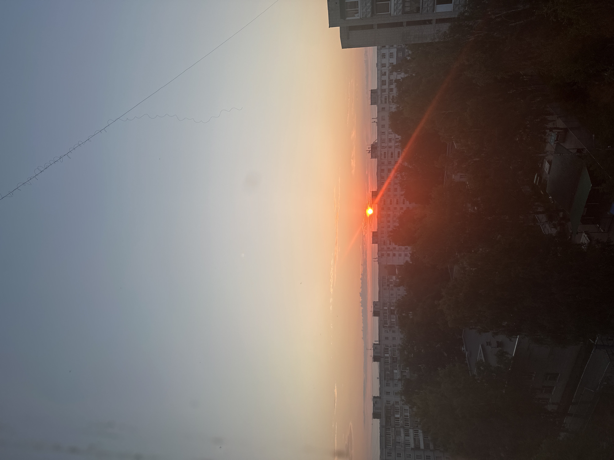 A wide-angle, eye-level shot captures a sunrise over a residential area. The sky transitions from a pale blue at the top to a soft orange and pink gradient near the horizon. A thin, dark wire with a wavy pattern stretches diagonally across the upper portion of the sky. In the middle ground, a line of multi-story apartment buildings, characteristic of Soviet-era architecture, stretches across the frame. The buildings are light gray with numerous windows. A low-lying layer of fog or haze obscures the ground beyond the buildings. In the foreground, dense green trees partially obscure the lower levels of the buildings. The sun, a bright orange orb, is visible just above the horizon, casting a warm glow and a lens flare across the scene. The overall impression is a tranquil urban landscape at dawn. The specific location is the city of Bakhchisarai, Russia.