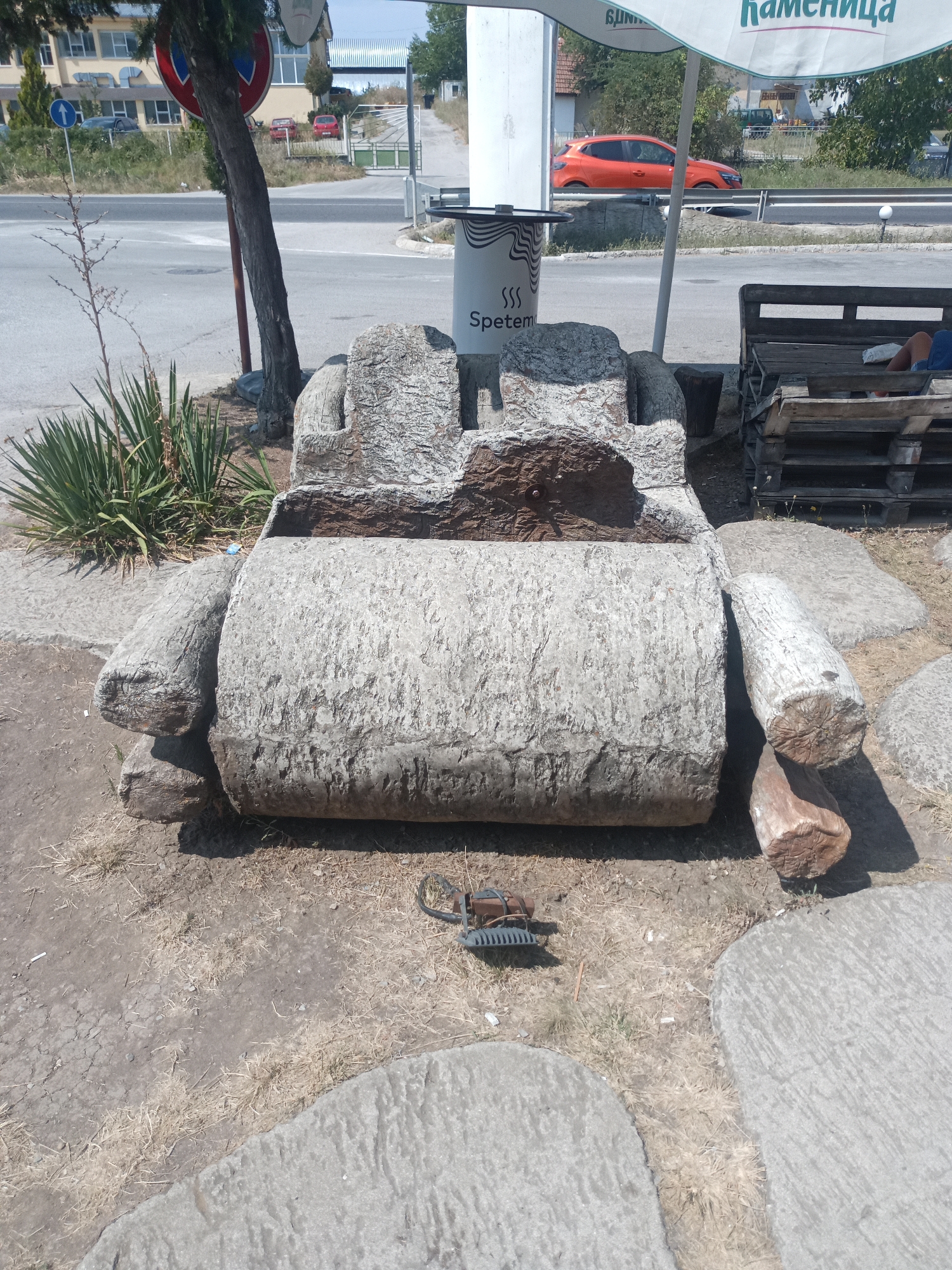 The image shows an outdoor scene in Omurtag, Bulgaria. In the foreground, a large, rough-hewn stone sculpture resembling an armchair or couch is prominently displayed. The sculpture is made of light-colored, textured stone and has a dark brown stone insert or cushioning. To the left and right of the main sculpture, there are large, cylindrical stone elements that appear to be part of the artwork or its base. Further to the right, a wooden pallet bench with a person seated on it is visible, suggesting this may be an outdoor seating area or a roadside attraction. In the background, a road with cars, buildings, trees, and a promotional umbrella are visible, indicating an urban or semi-urban environment. The ground is a mix of dirt, dry grass, and large stone slabs. A small, metal object resembling a garden tool lies on the ground in front of the sculpture.