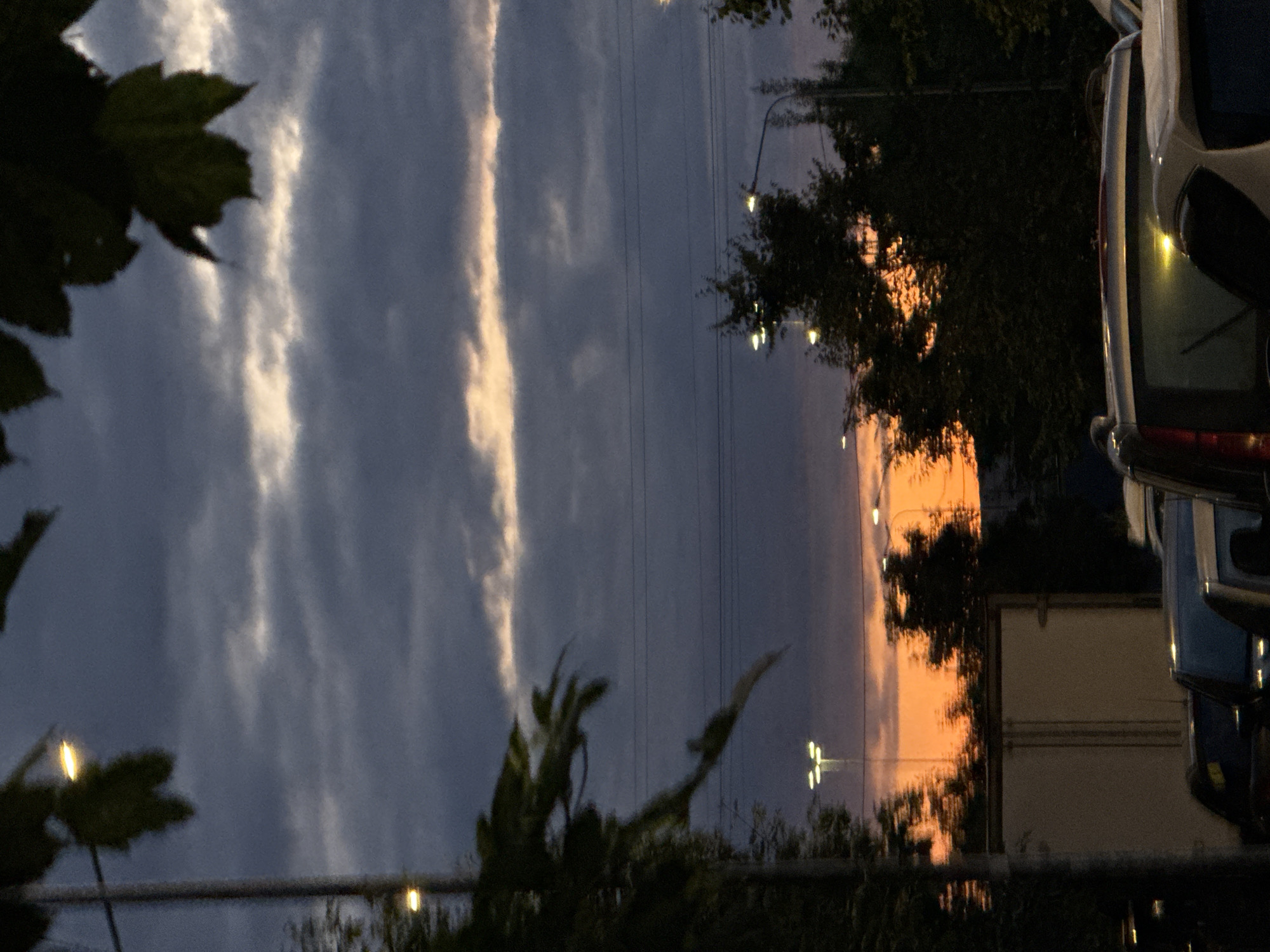This image captures a dusky scene in Shushary, Russia. The sky is a dramatic mix of deep blues and grays, with a bright orange band visible near the horizon, indicating sunset or sunrise. Several dark clouds are scattered across the sky, and faint horizontal lines, likely power lines, are visible. In the foreground, silhouetted trees and streetlights punctuate the darkening landscape. At the bottom of the frame, the tops of several vehicles are visible, suggesting a road or parking area. The image has a somewhat blurred or soft focus, contributing to the atmospheric quality of the twilight setting.