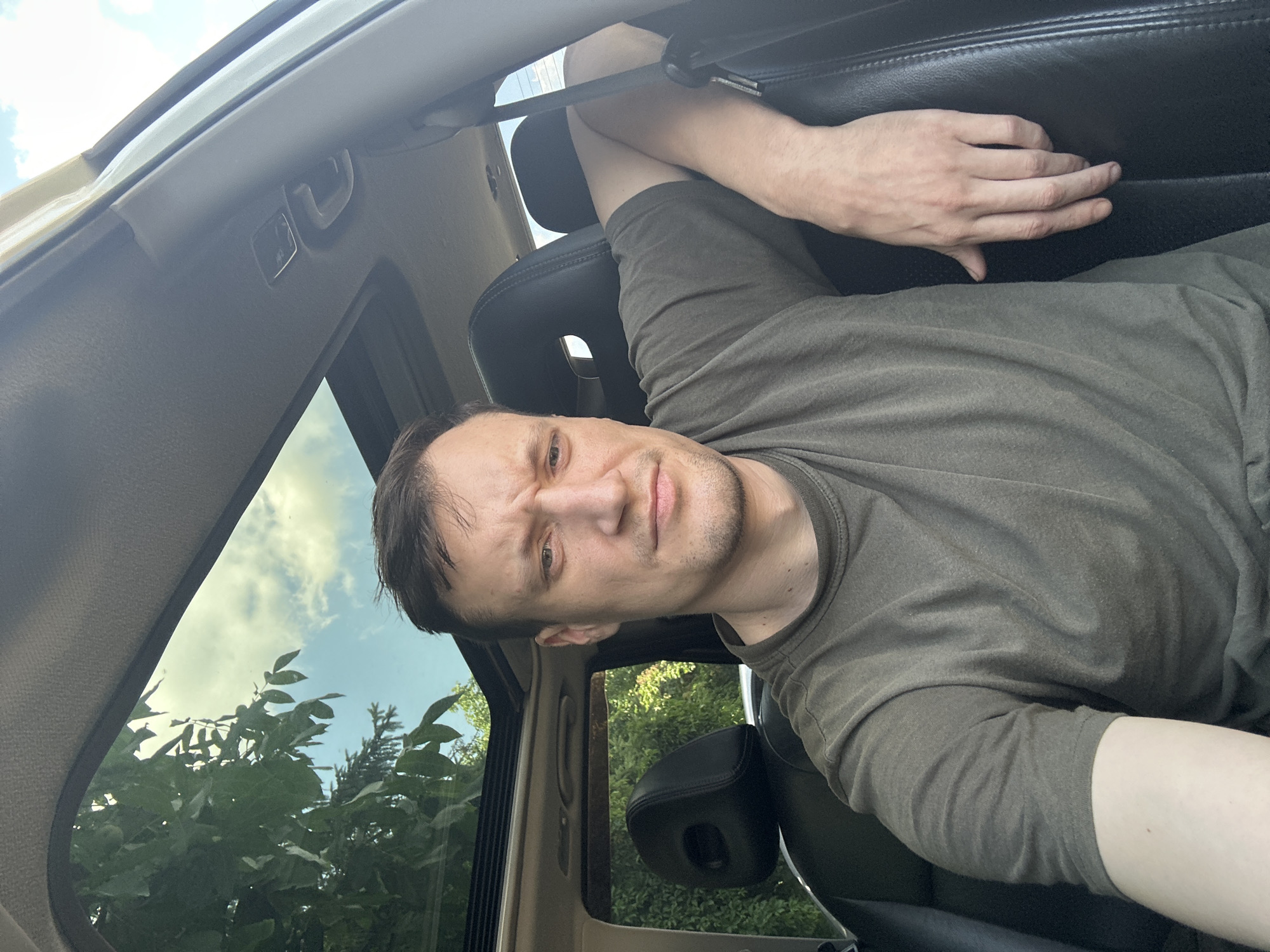 In Shatava, Ukraine, a man with short, dark hair and a light complexion is seated inside a vehicle. He is wearing a dark green t-shirt and has his right arm resting on the leather seat. The man is looking directly at the camera with a slightly furrowed brow. The sunroof of the car is open, revealing a sky with scattered clouds and green foliage. The interior of the car appears to be a dark color, possibly black leather for the seats.