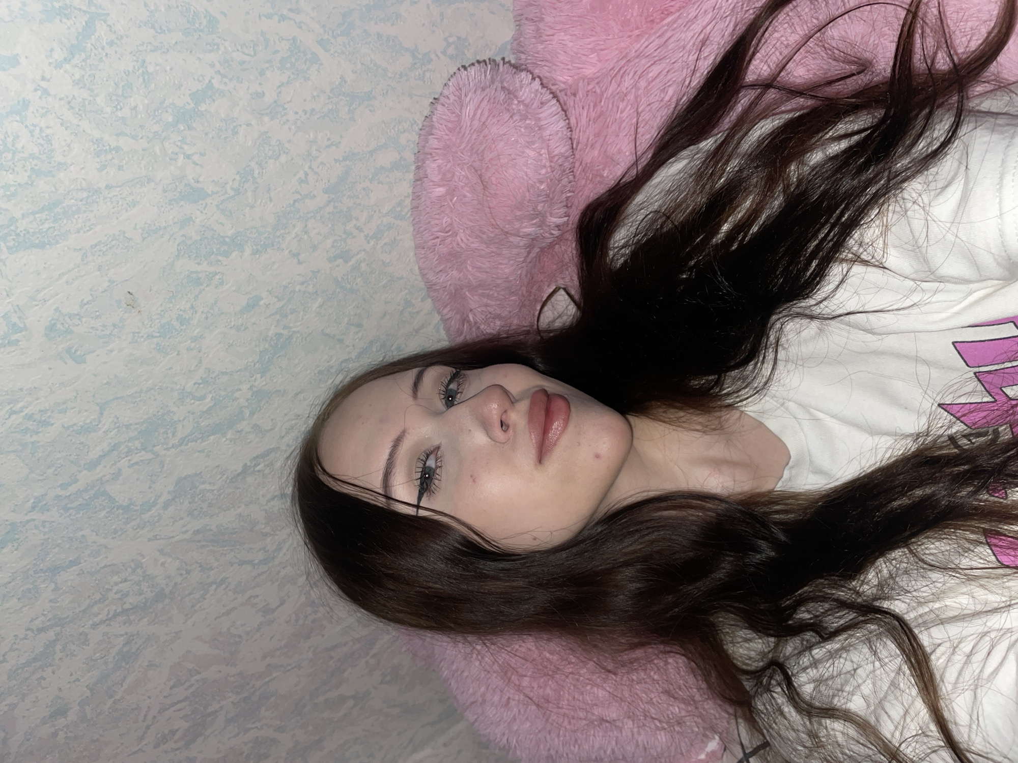 The image depicts a young woman with long, dark brown hair, wearing a white t-shirt. She is lying down, propped up against a large, fluffy pink object, possibly a pillow or stuffed animal. The background appears to be a wall with a textured, patterned wallpaper in shades of light blue and white. The lighting suggests an indoor setting, likely a bedroom. There is no information in the image that would allow for the determination of its location within Votkinsk, Russia.